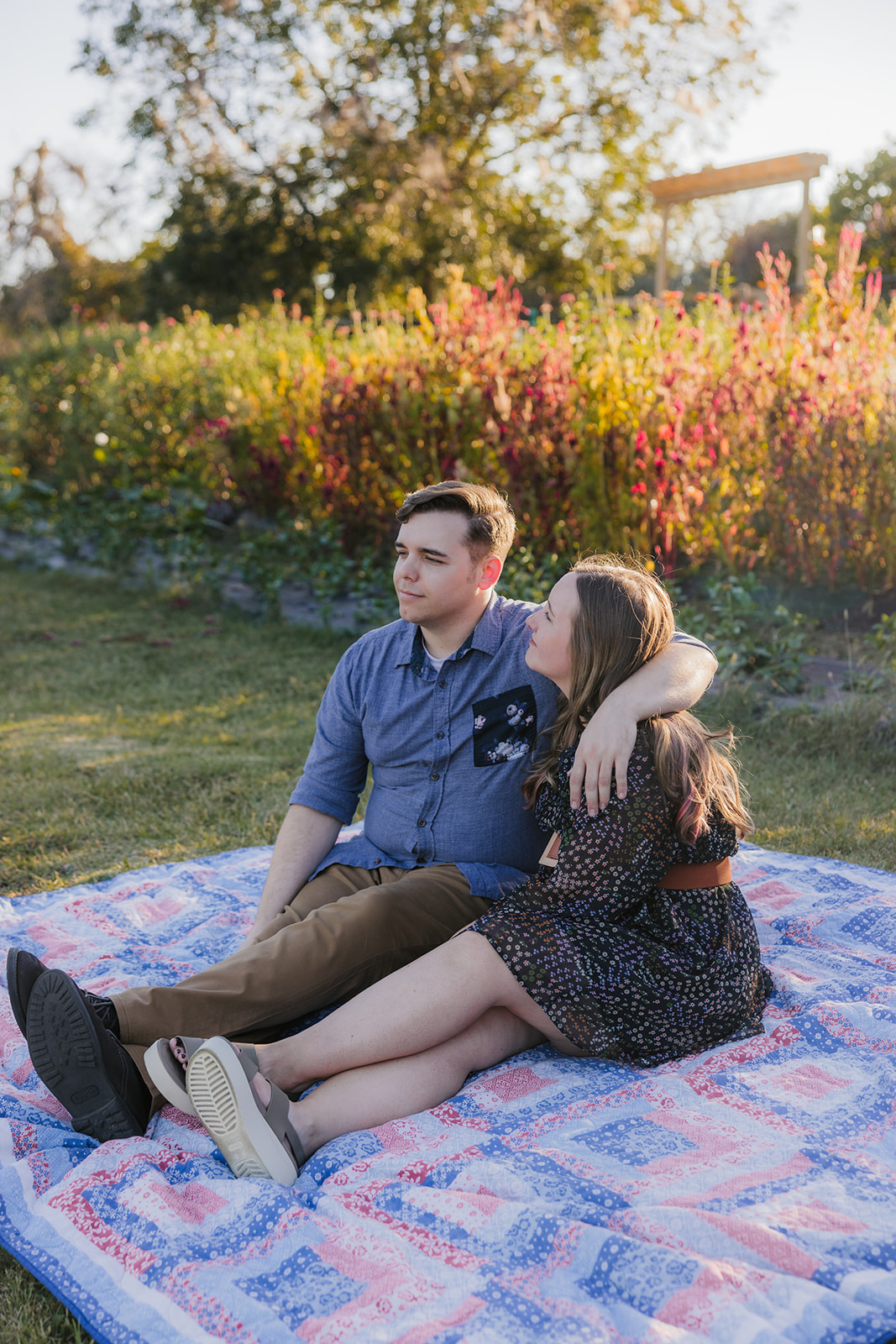 A couple sits on a blanket in a garden, surrounded by plants and trees, enjoying a sunny day.