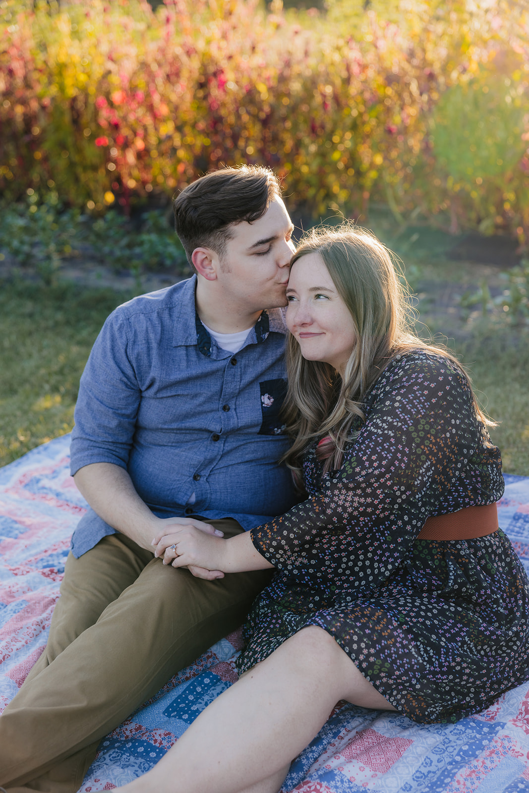 A couple sits on a blanket in a garden, surrounded by plants and trees, enjoying a sunny day.