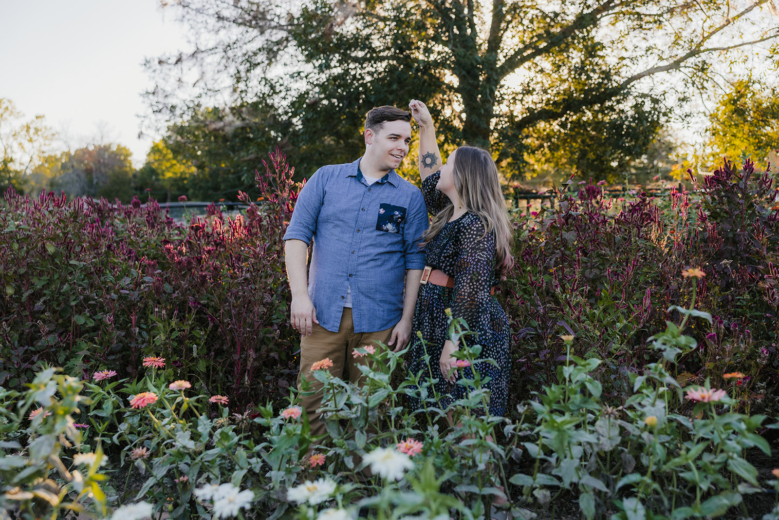 A couple sits on a blanket in a garden, surrounded by plants and trees, enjoying a sunny day.