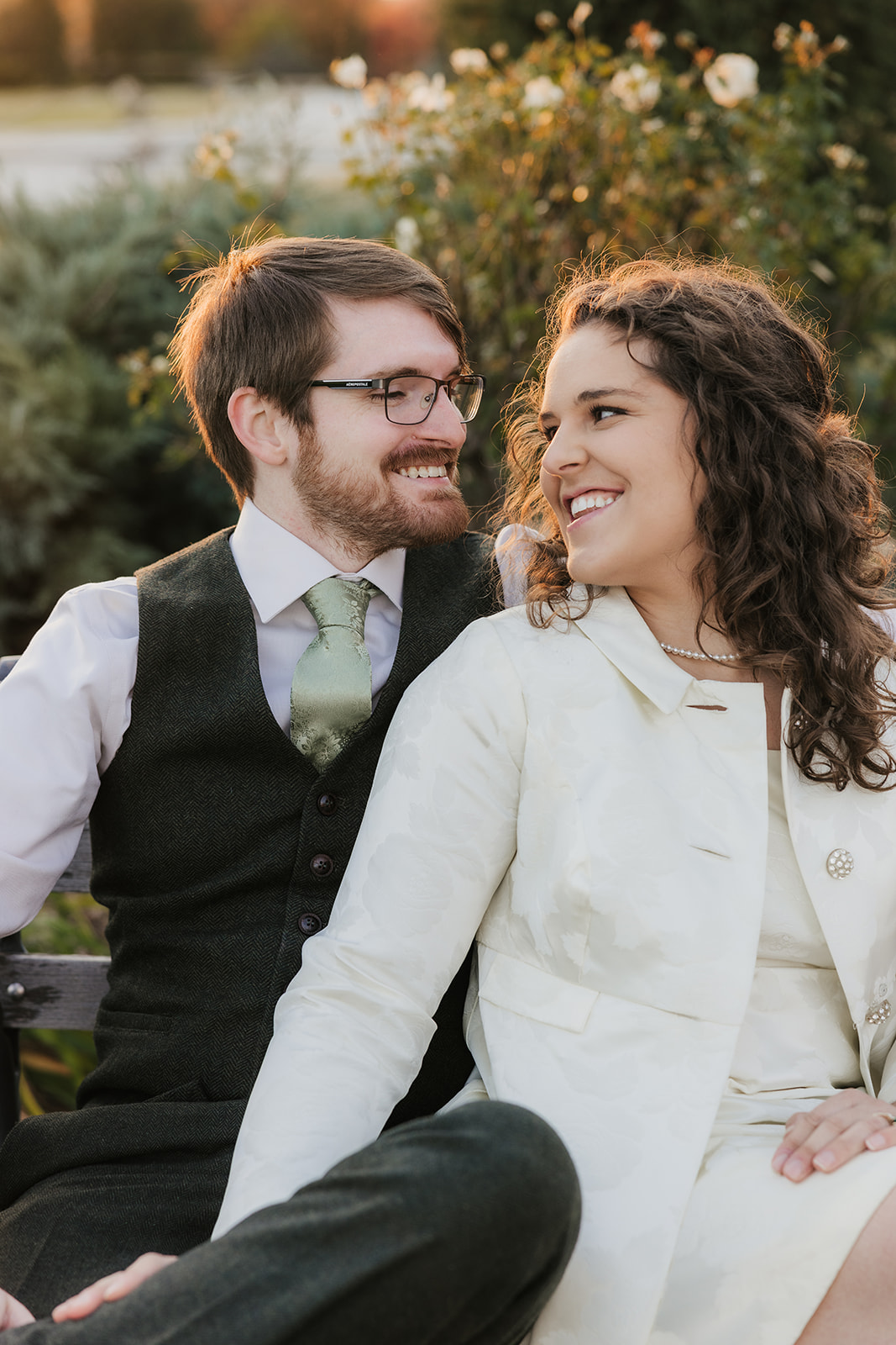 A couple sitting on a bench in a garden, looking at each other and smiling. The man wears a vest and tie, and the woman is in a light-colored coat for their Tulsa engagement photos