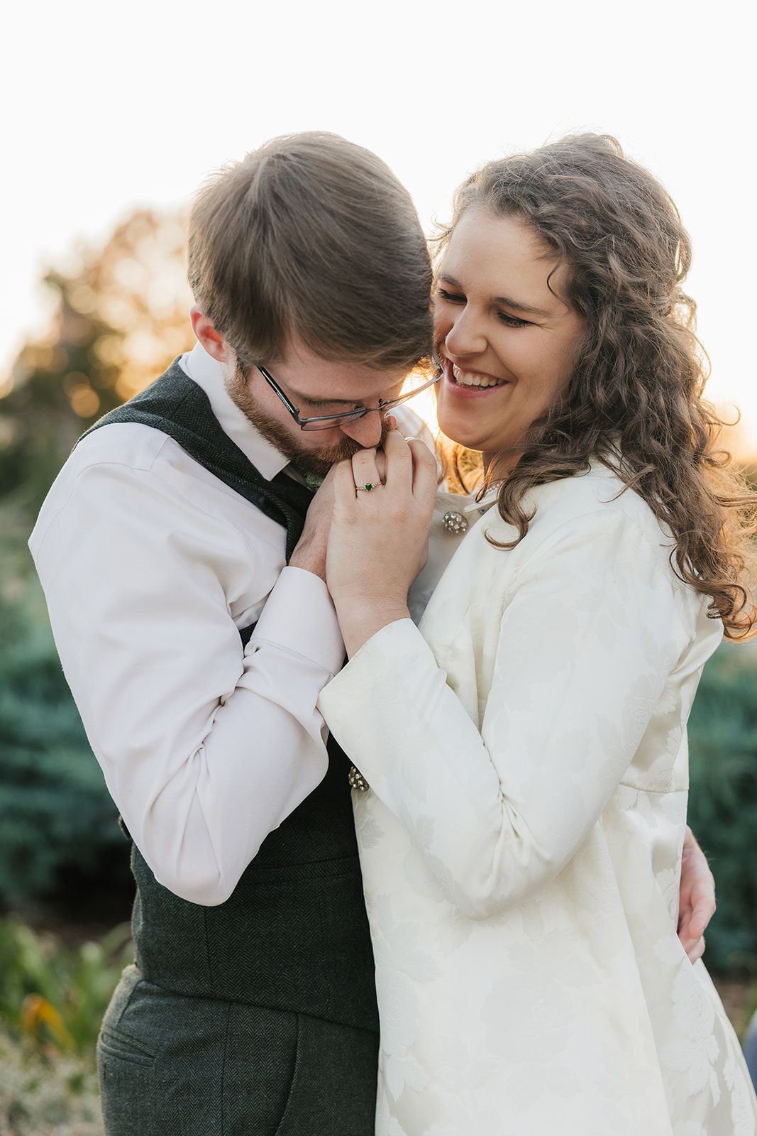 A couple sitting on a bench in a garden, looking at each other and smiling. The man wears a vest and tie, and the woman is in a light-colored coat for their Tulsa engagement photos