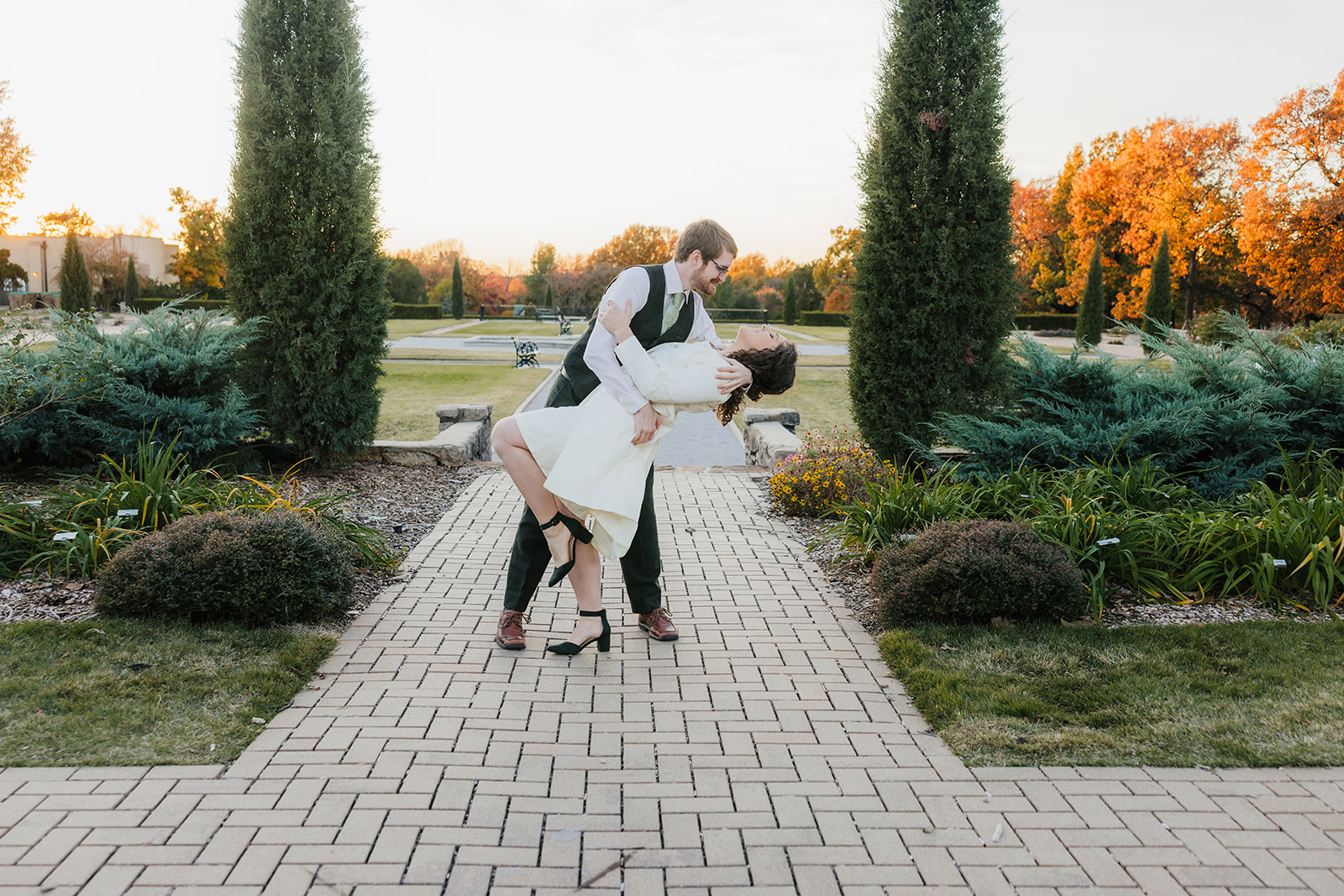 A person in a suit dips a person in a white dress on a brick path surrounded by tall trees for their tulsa engagement photos