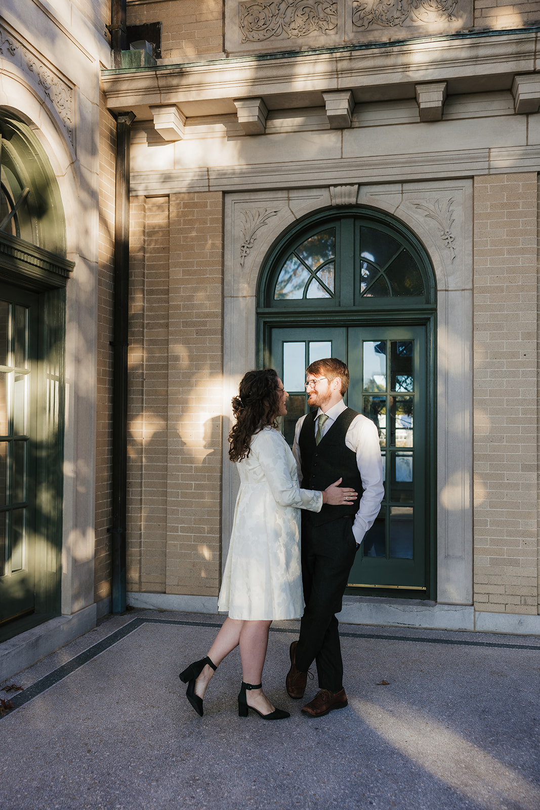 A couple stands closely together, smiling, in front of an ornate building with green arched windows at woodward park for their tulsa engagement photos