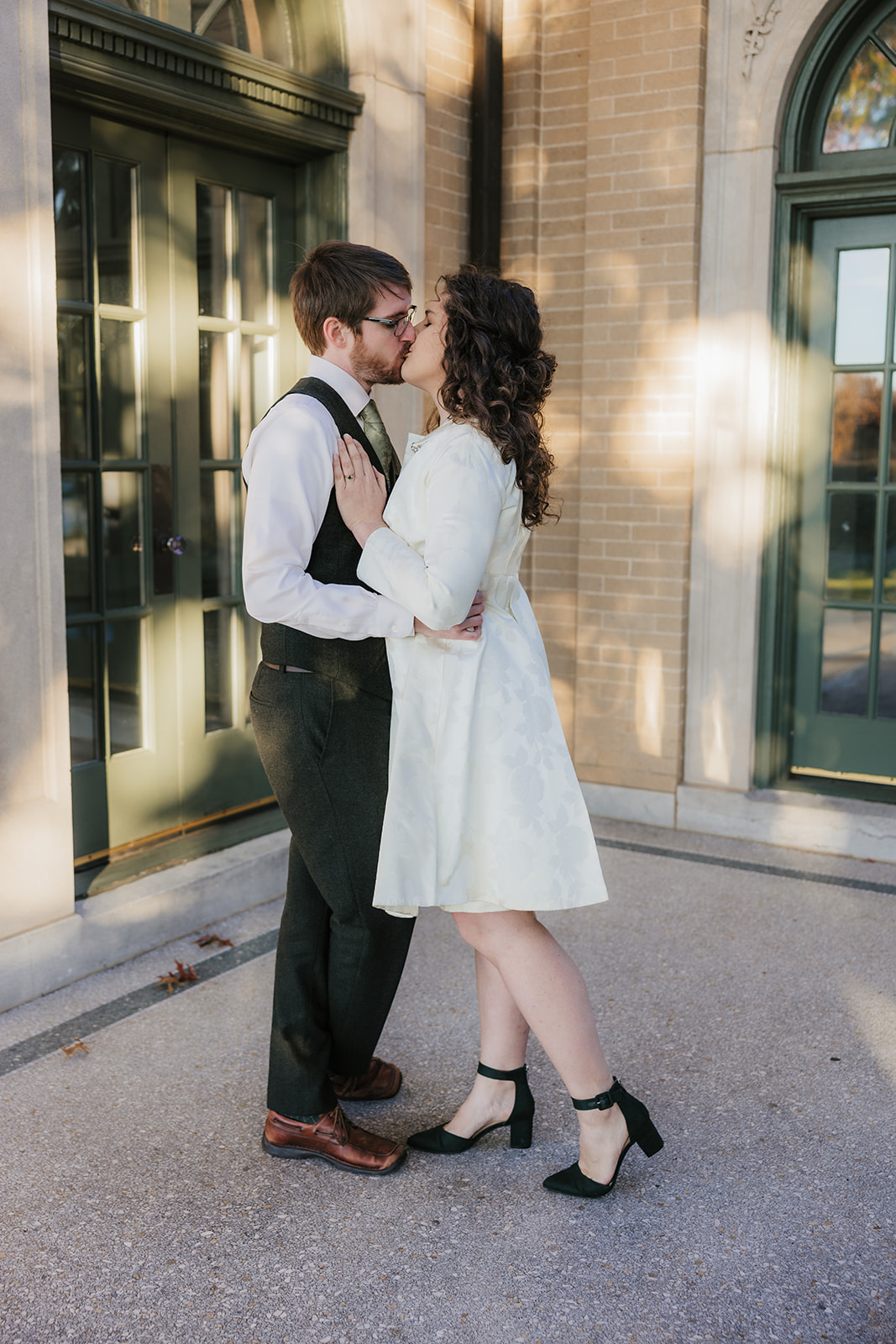 A couple stands closely together, smiling, in front of an ornate building with green arched windows at woodward park for their tulsa engagement photos 