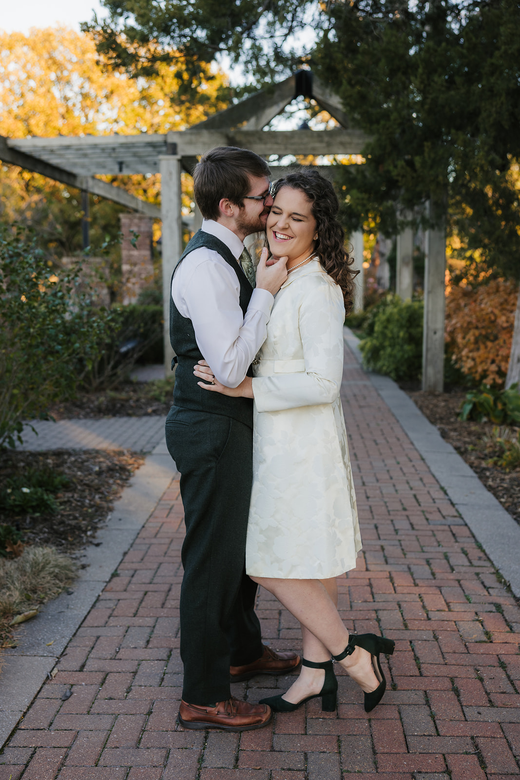 A couple dressed in formal attire stands on a brick pathway in a garden. The man is whispering to the woman, who is smiling.