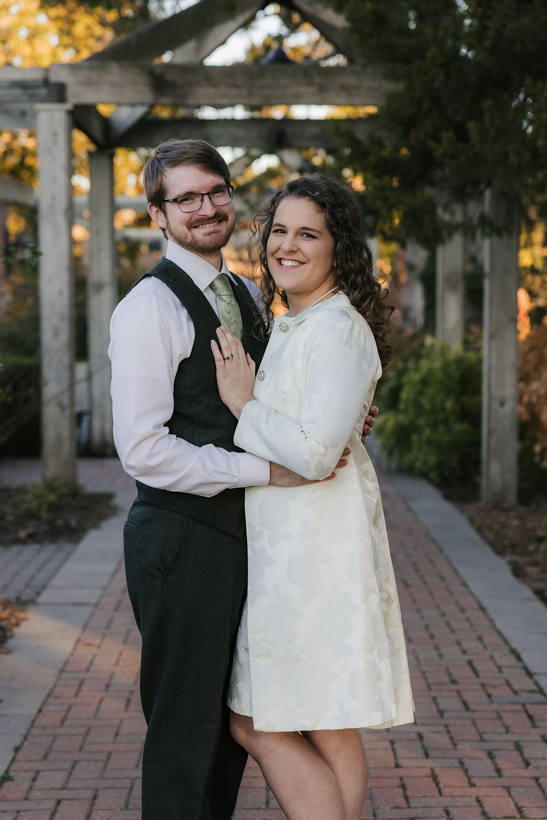 A couple dressed in formal attire stands on a brick pathway in a garden. The man is whispering to the woman, who is smiling.