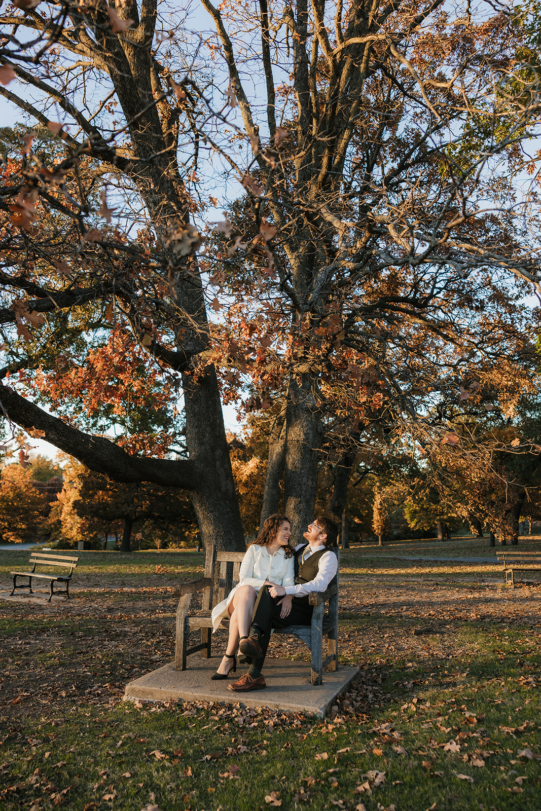 A couple sits on a park bench under a large tree with autumn leaves, surrounded by fallen leaves and late afternoon sunlight.