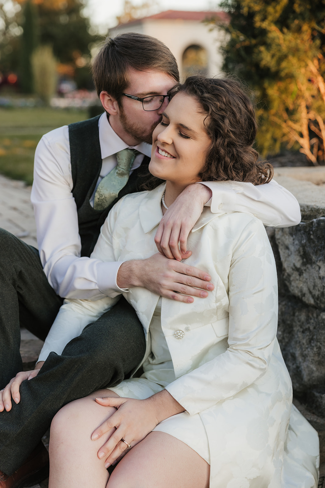 A couple sitting on a bench in a garden, looking at each other and smiling. The man wears a vest and tie, and the woman is in a light-colored coat for their Tulsa engagement photos