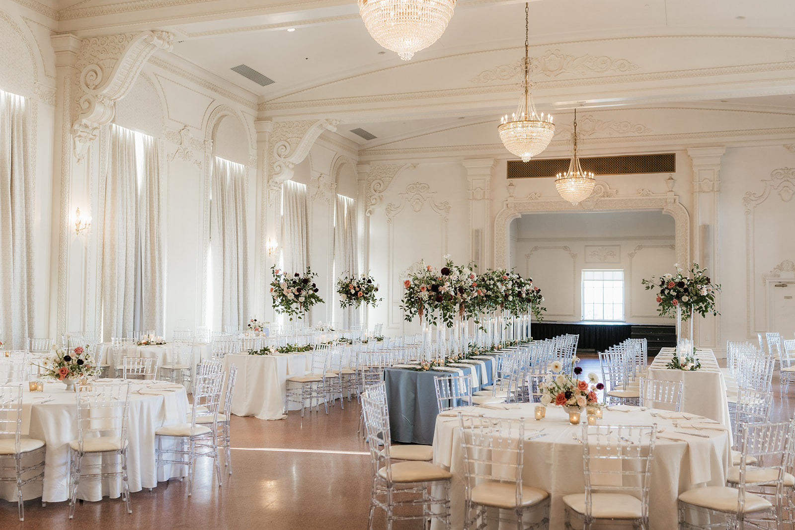 Elegant banquet hall with round tables draped in white linens, clear chairs, and floral centerpieces. Large chandeliers hang from the high ceiling at the mayo hotel for a wedding