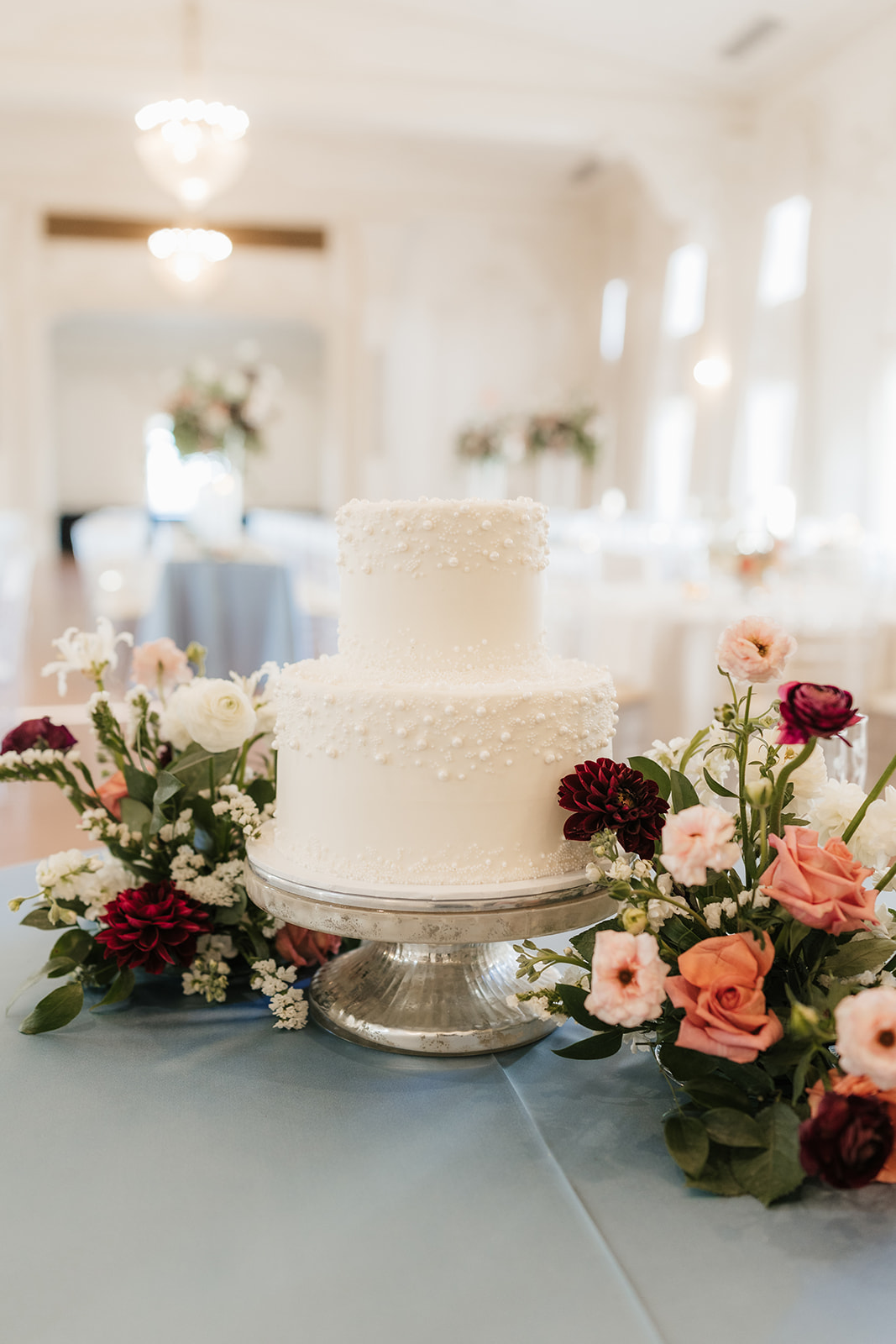 A two-tier white cake on a silver stand is surrounded by pink and red flowers on a blue tablecloth in a bright room.