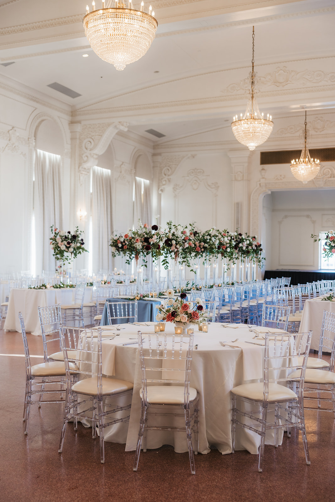 Elegant banquet hall with round tables draped in white linens, clear chairs, and floral centerpieces. Large chandeliers hang from the high ceiling at the mayo hotel for a wedding