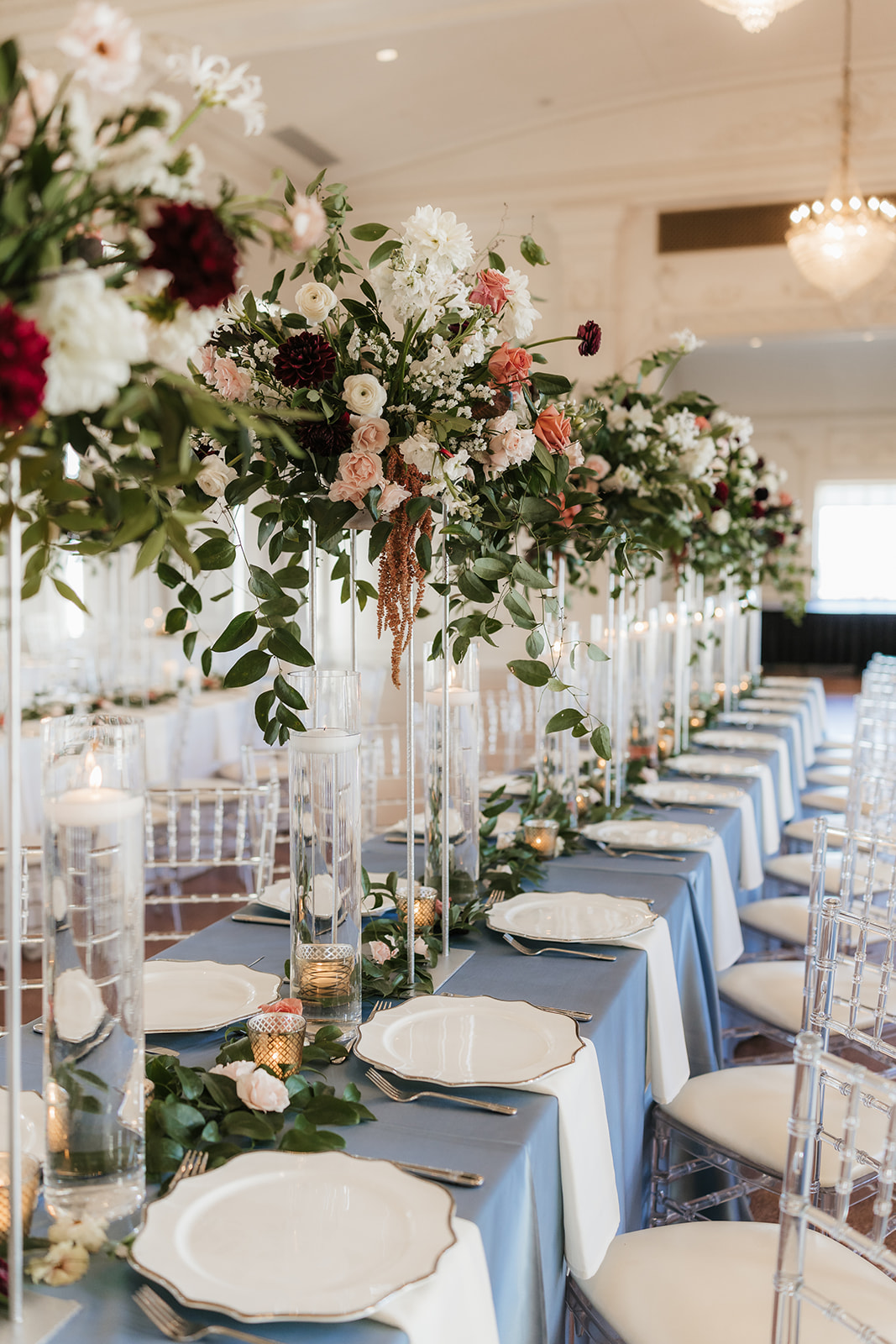 Elegant banquet hall with round tables draped in white linens, clear chairs, and floral centerpieces. Large chandeliers hang from the high ceiling at the mayo hotel for a wedding