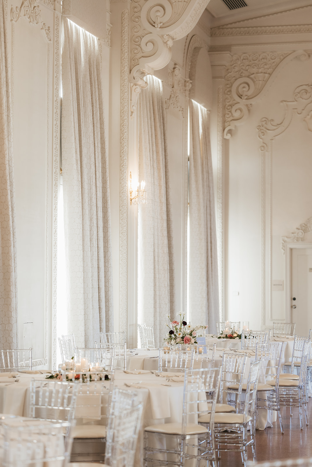 Elegant banquet hall with round tables draped in white linens, clear chairs, and floral centerpieces. Large chandeliers hang from the high ceiling at the mayo hotel for a wedding