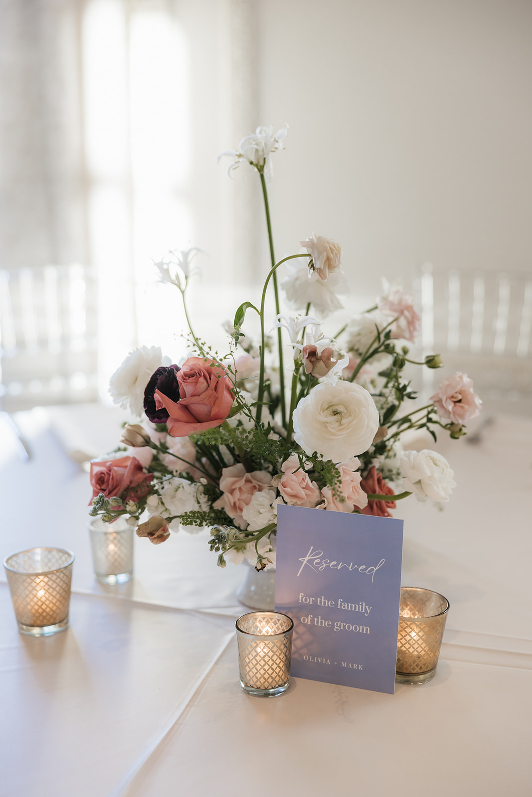 Floral centerpiece with pink and white flowers on a table, surrounded by small lit candles. A card with text is in front of the arrangement.