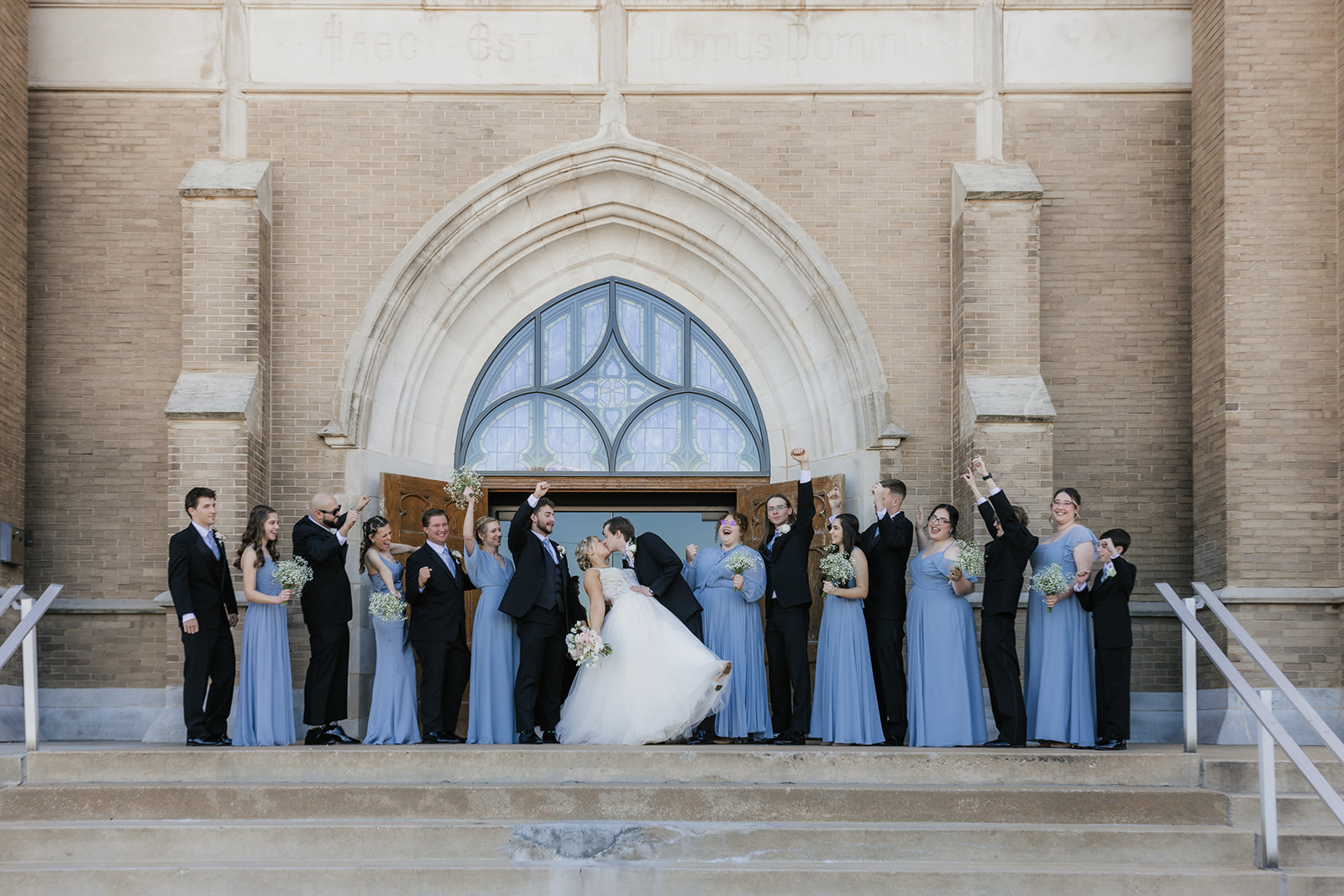 A bride and groom stand on church steps, surrounded by a cheering wedding party in blue dresses and black suits at the holy family cathedral 