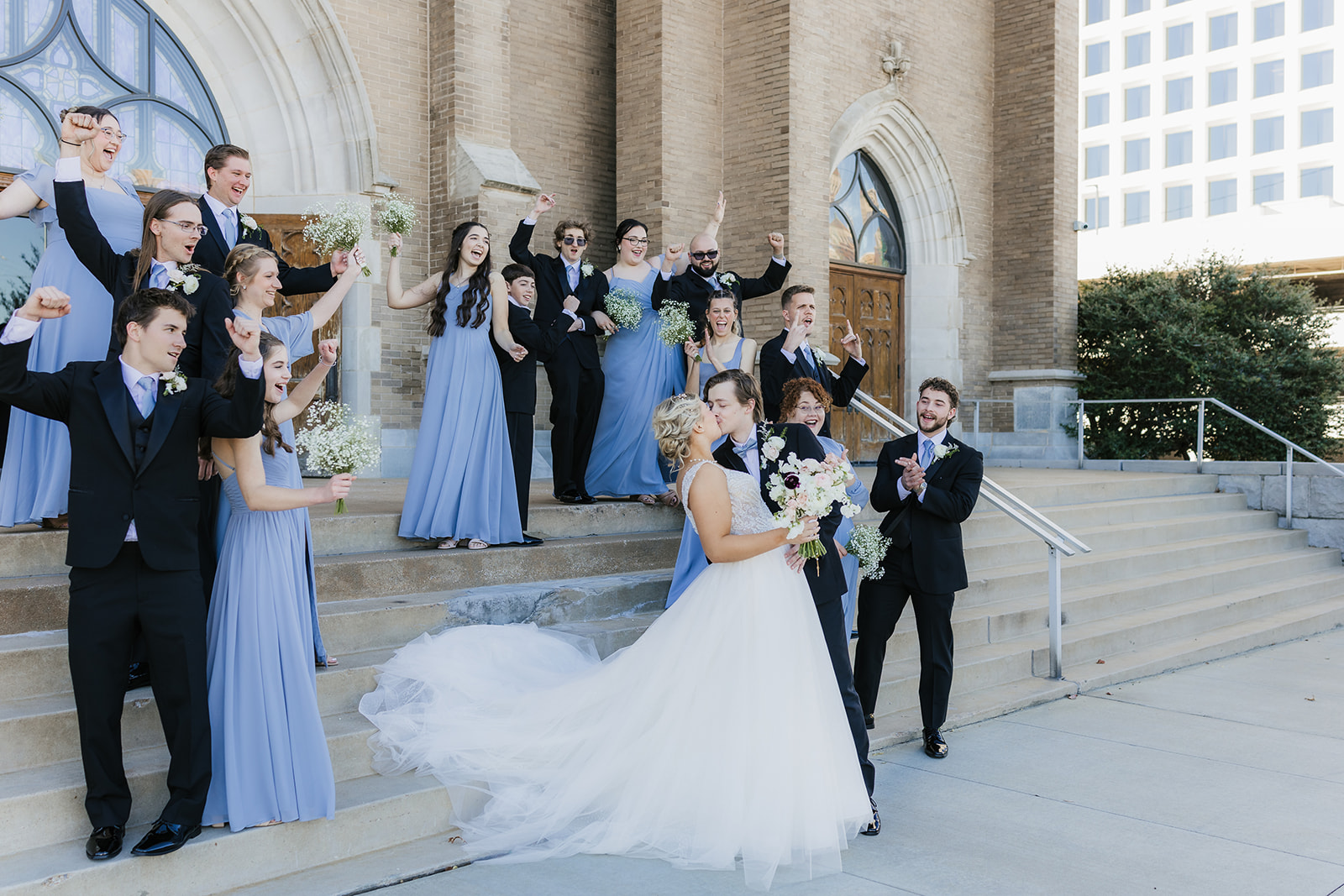 A bride and groom stand on church steps, surrounded by a cheering wedding party in blue dresses and black suits at the holy family cathedral 