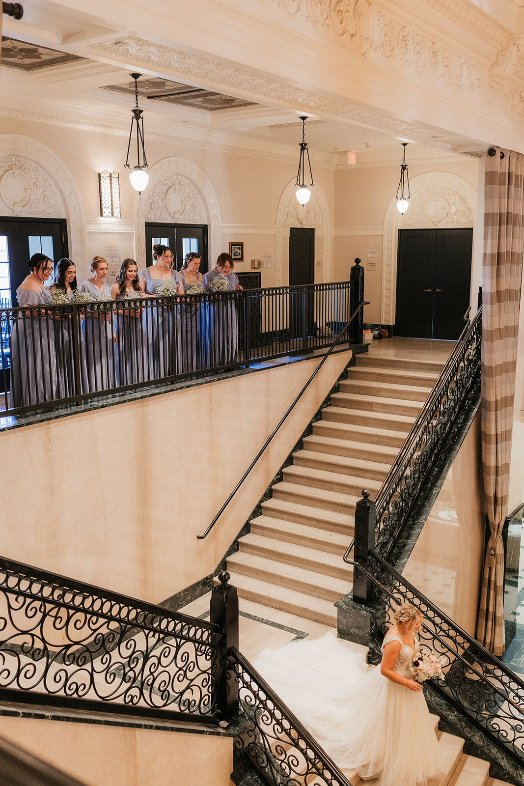 Bride walks up a grand staircase while a group of people observes from the balcony above in a formal setting.