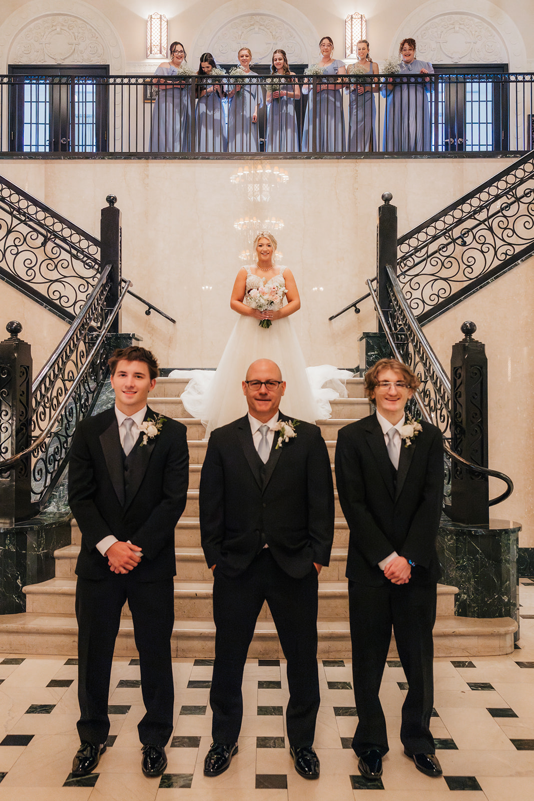 Three men in suits stand on the stairway of an elegant building, while a woman in a white dress poses at the top of the stairs.