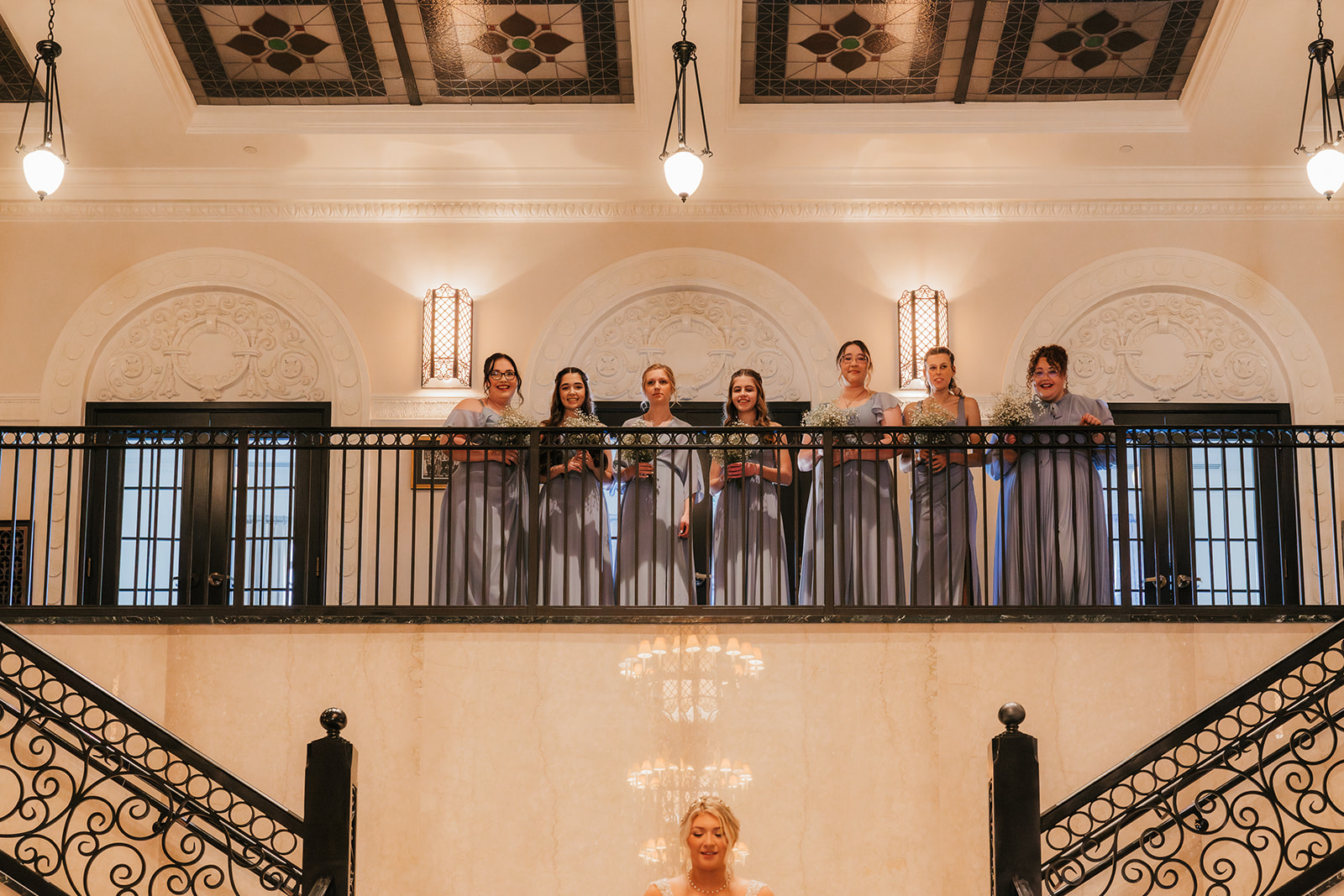 Bride walks up a grand staircase while a group of people observes from the balcony above in a formal setting.