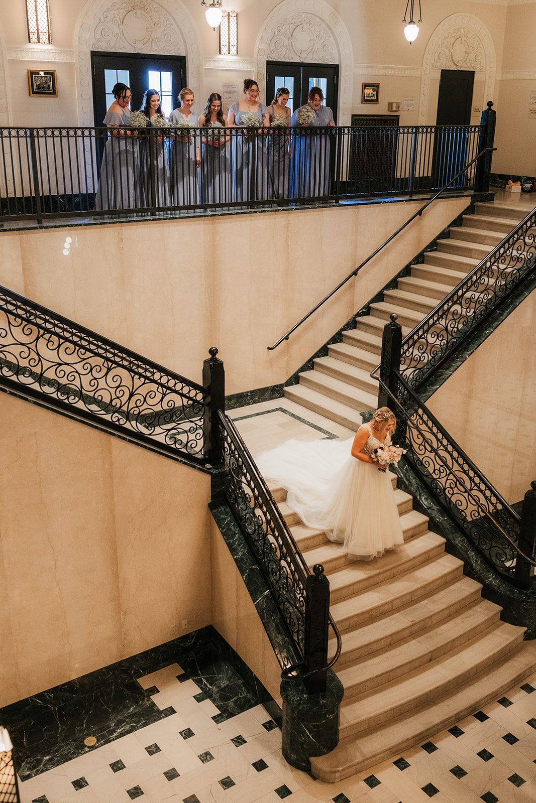 Bride walks up a grand staircase while a group of people observes from the balcony above in a formal setting.