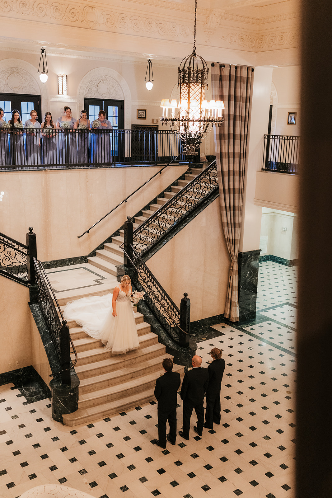 Three men in suits stand on the stairway of an elegant building, while a woman in a white dress poses at the top of the stairs.