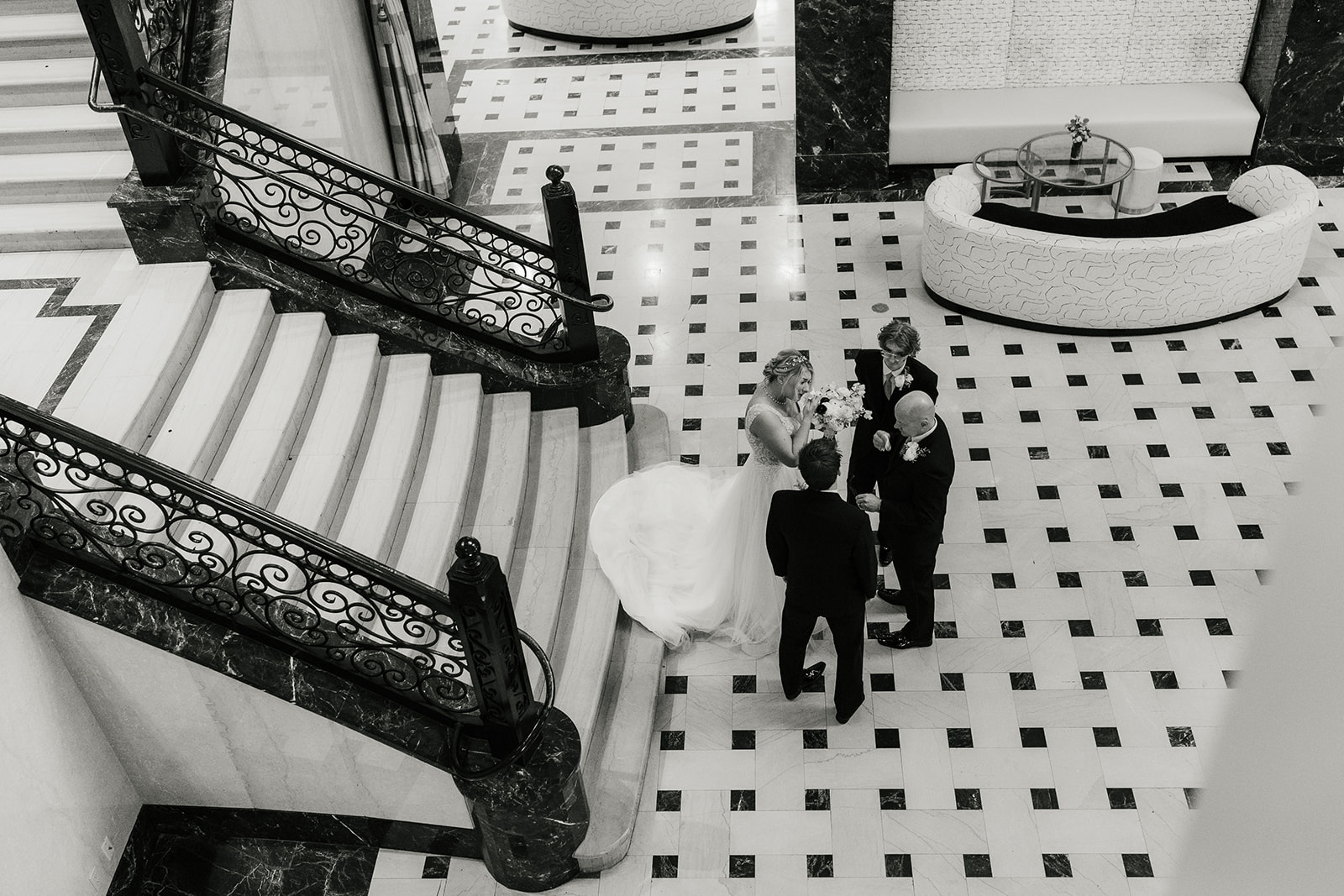 Three men in suits stand on the stairway of an elegant building, while a woman in a white dress poses at the top of the stairs.