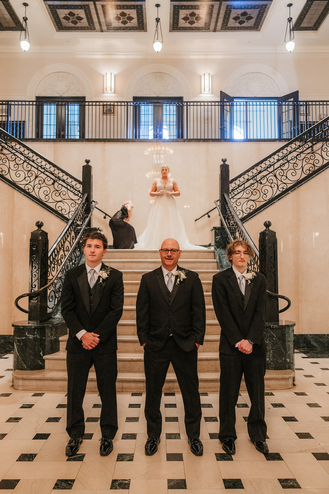 Three men in suits stand on the stairway of an elegant building, while a woman in a white dress poses at the top of the stairs.