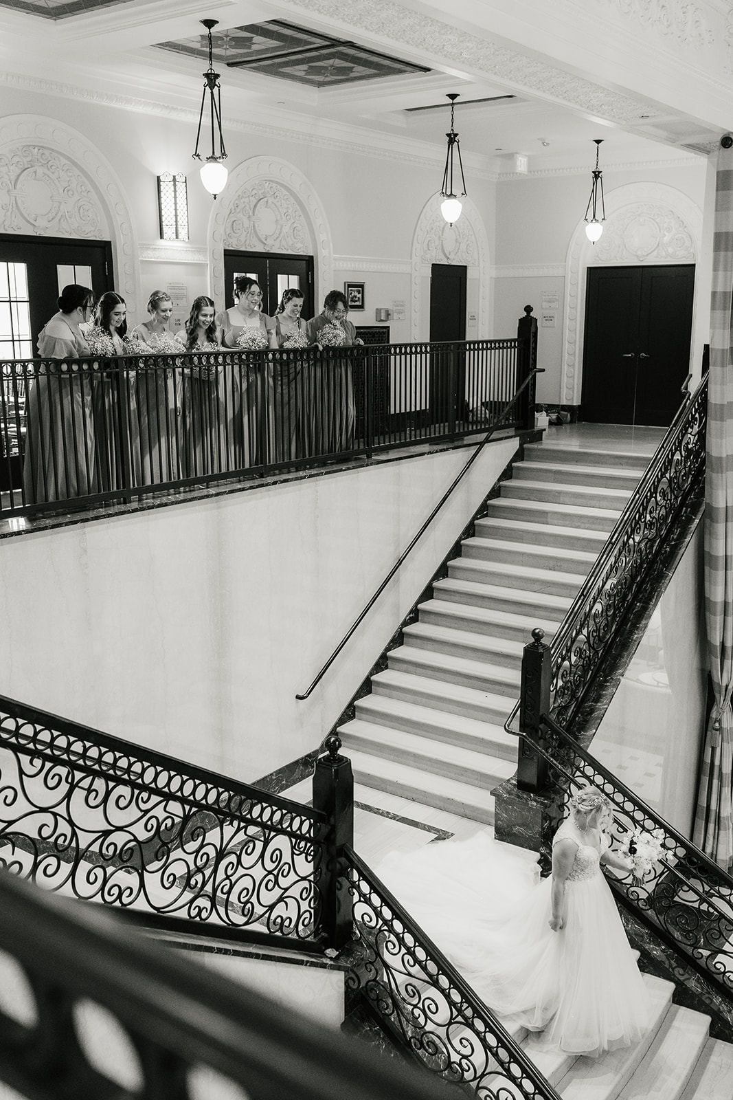 Bride walks up a grand staircase while a group of people observes from the balcony above in a formal setting.