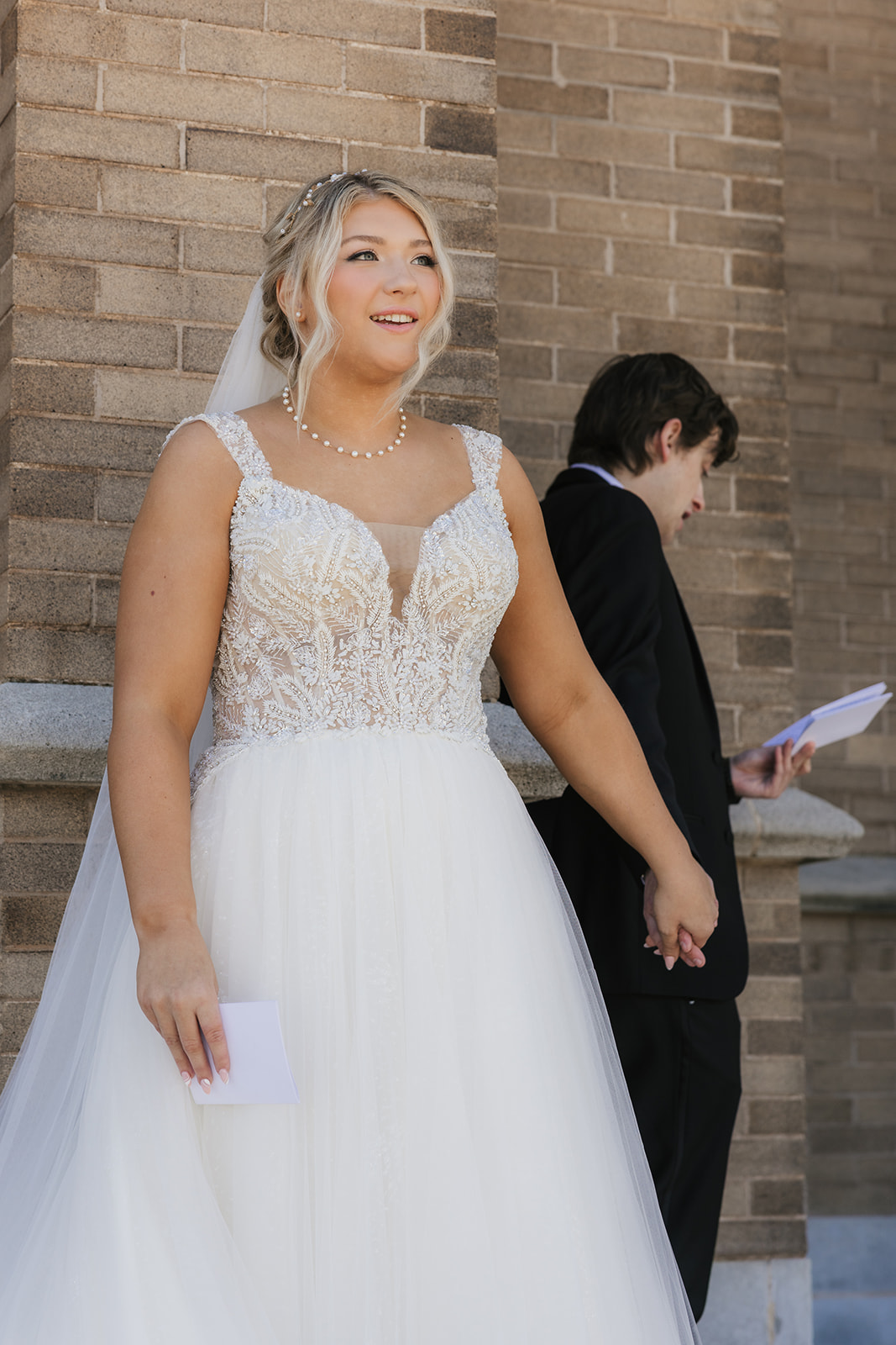 A bride and groom stand back-to-back outside a brick building, holding hands around a corner. The bride is reading from a card.