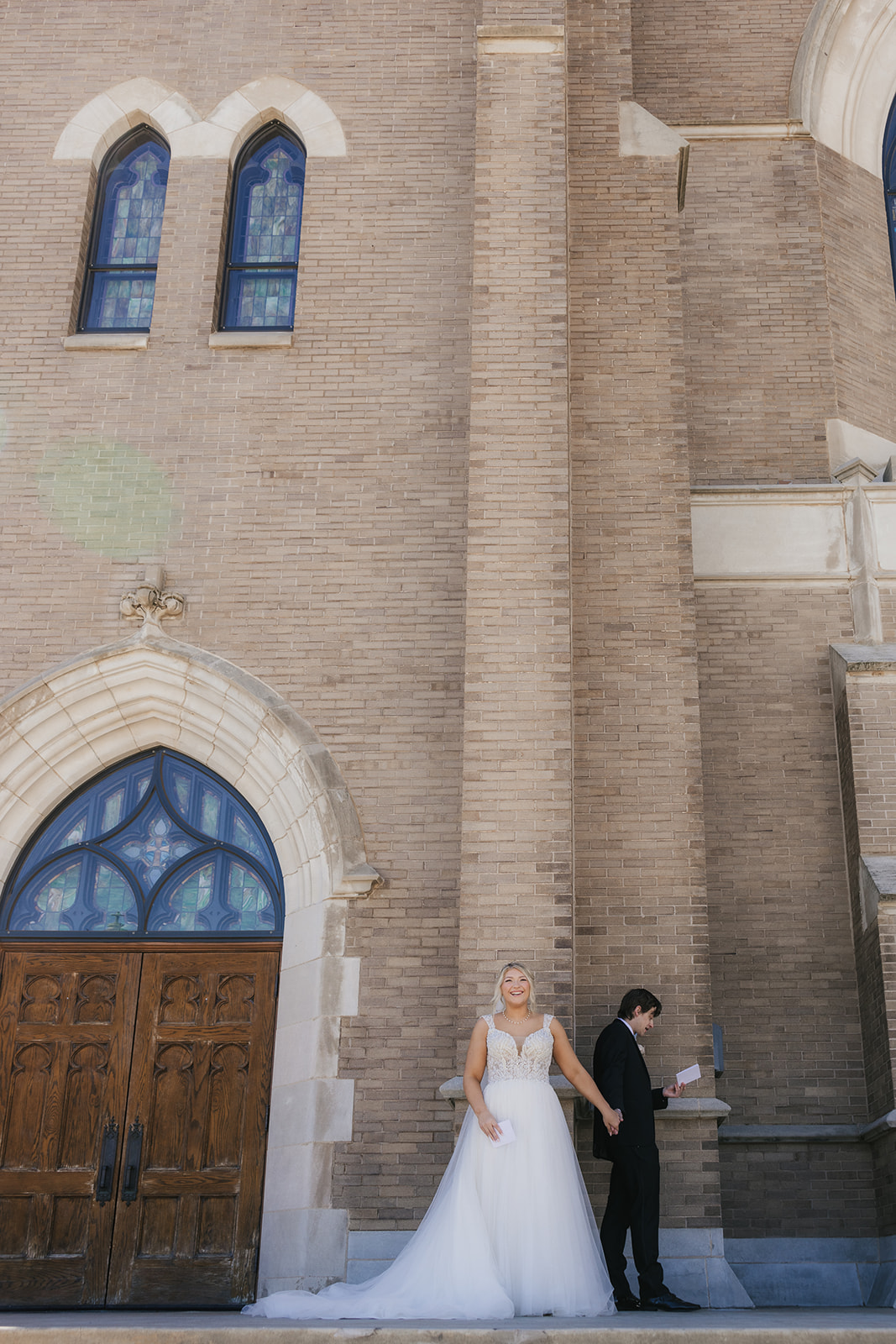 A bride and groom stand back-to-back outside a brick building, holding hands around a corner. The bride is reading from a card.