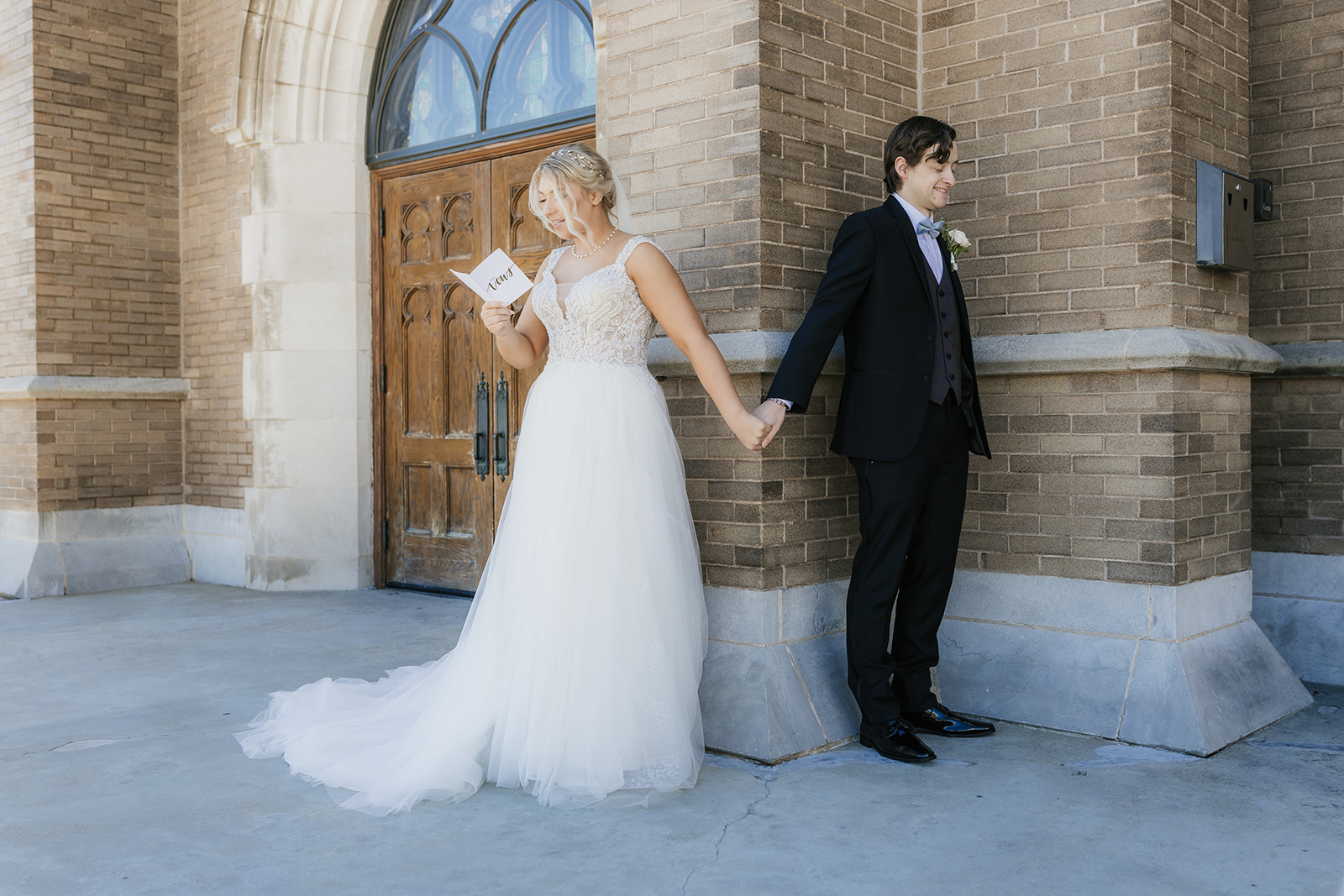 A bride and groom stand back-to-back outside a brick building, holding hands around a corner. The bride is reading from a card.