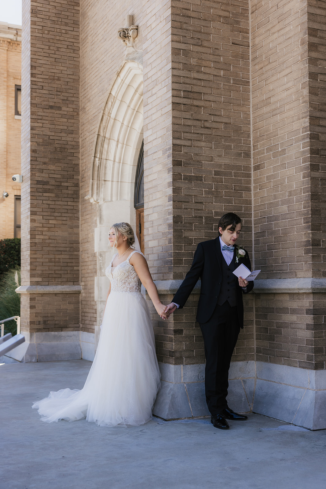 A bride and groom stand back-to-back outside a brick building, holding hands around a corner. The bride is reading from a card.