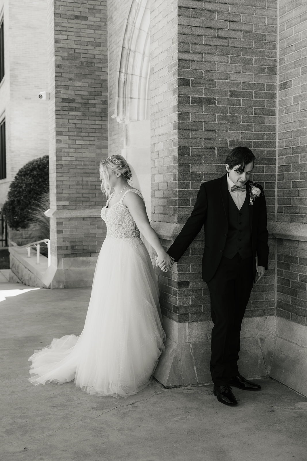 A bride and groom stand back-to-back outside a brick building, holding hands around a corner. The bride is reading from a card.