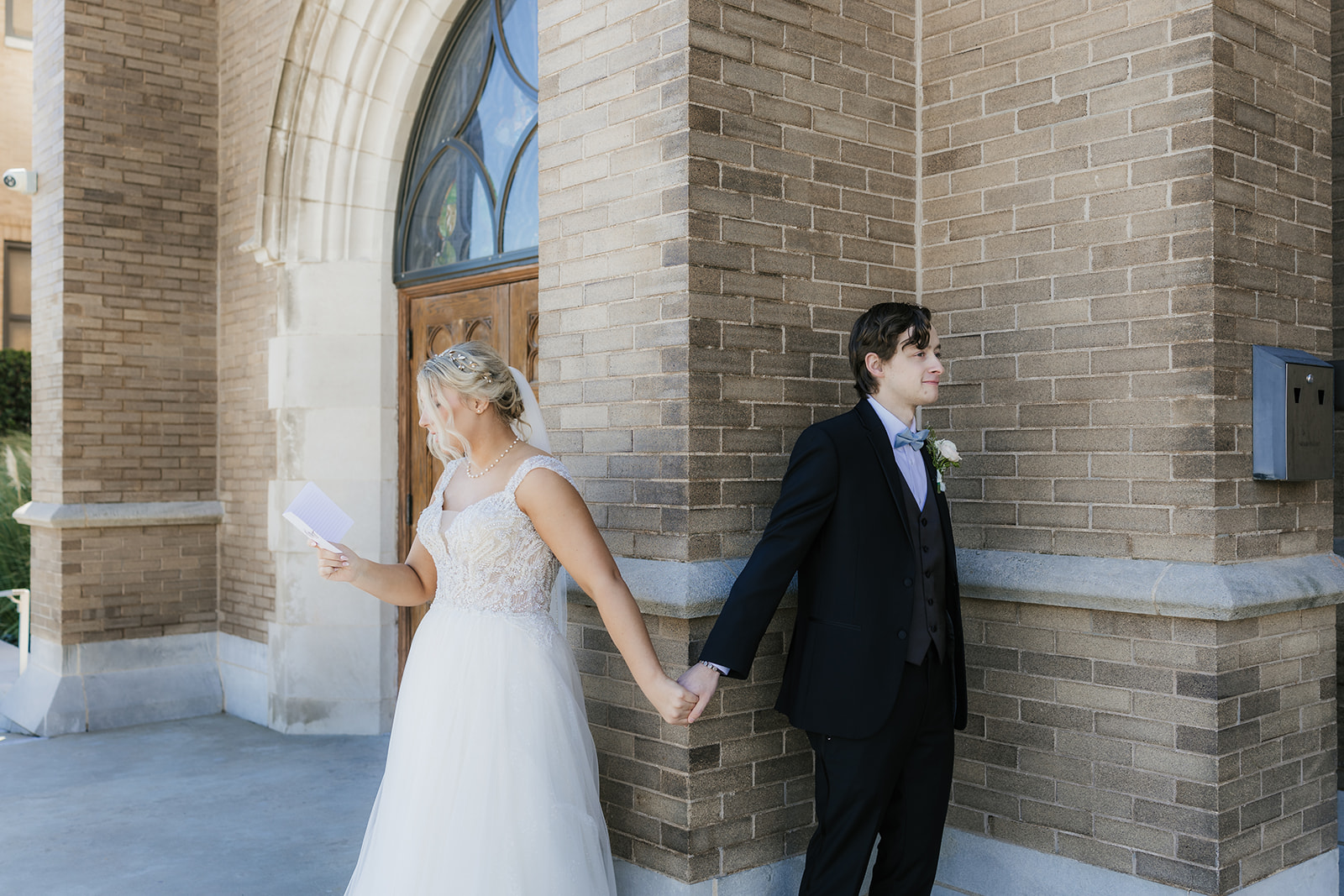 A bride and groom stand back-to-back outside a brick building, holding hands around a corner. The bride is reading from a card.