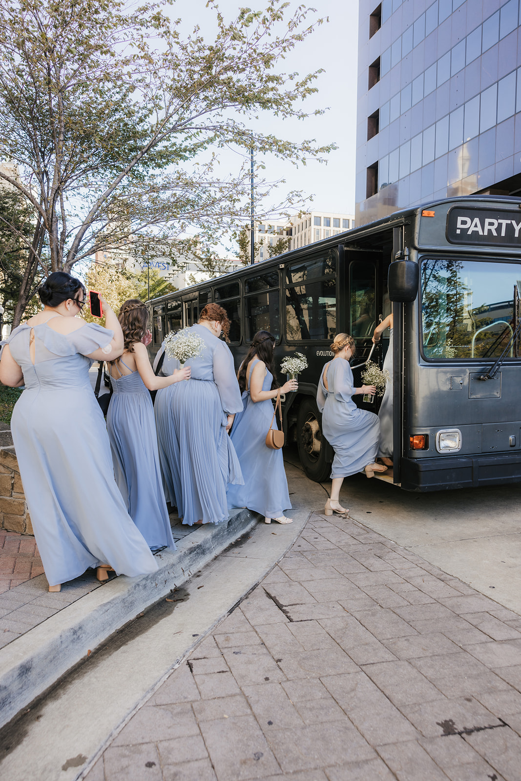 A group of women in matching blue dresses is boarding a black party bus in an urban setting.