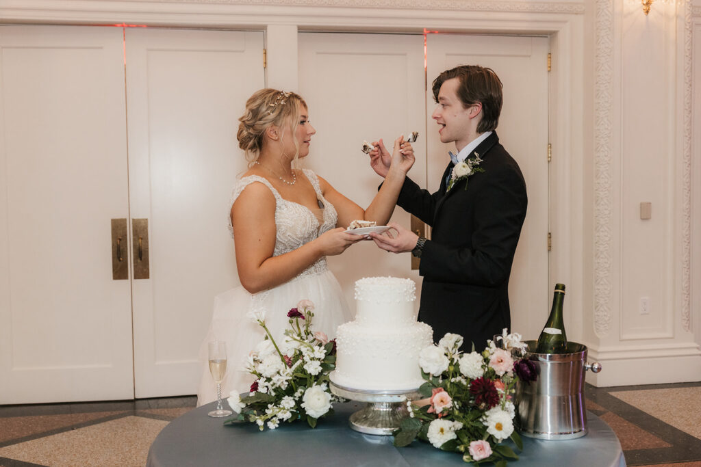 Bride and groom feed each other cake at their wedding reception, with a white cake and floral decoration on the table.