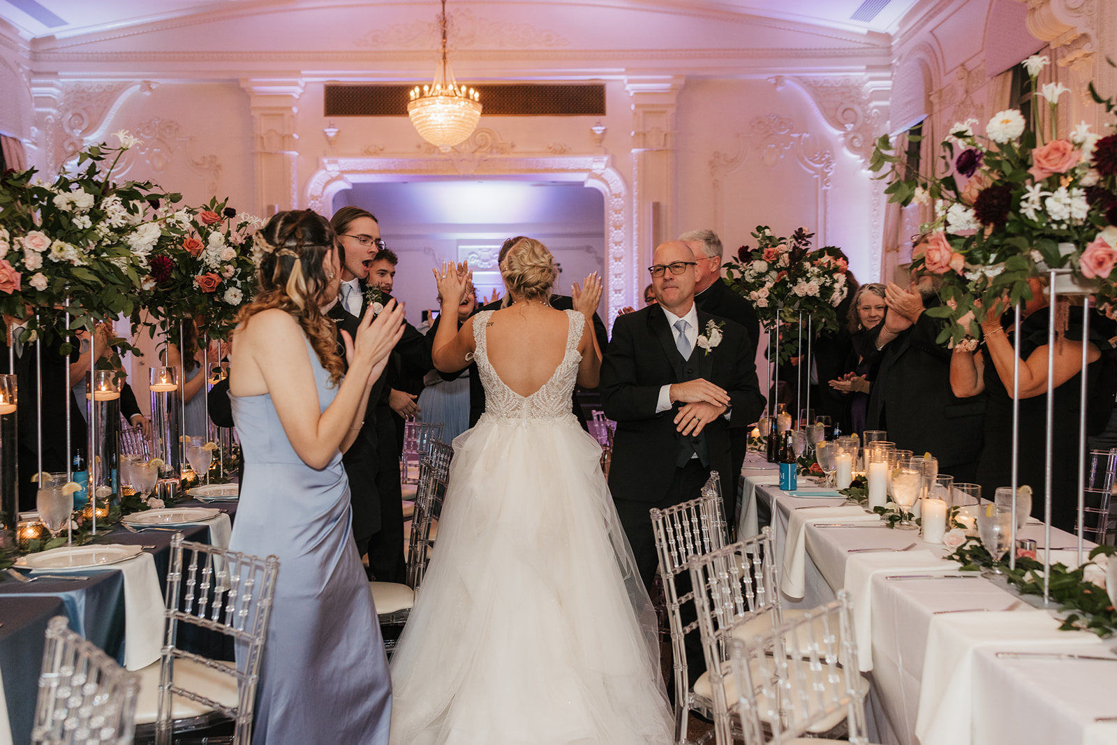 A bride and groom walk hand in hand through a decorated reception hall with guests on either side, wearing formal attire. Tables with candles and floral arrangements are visible.