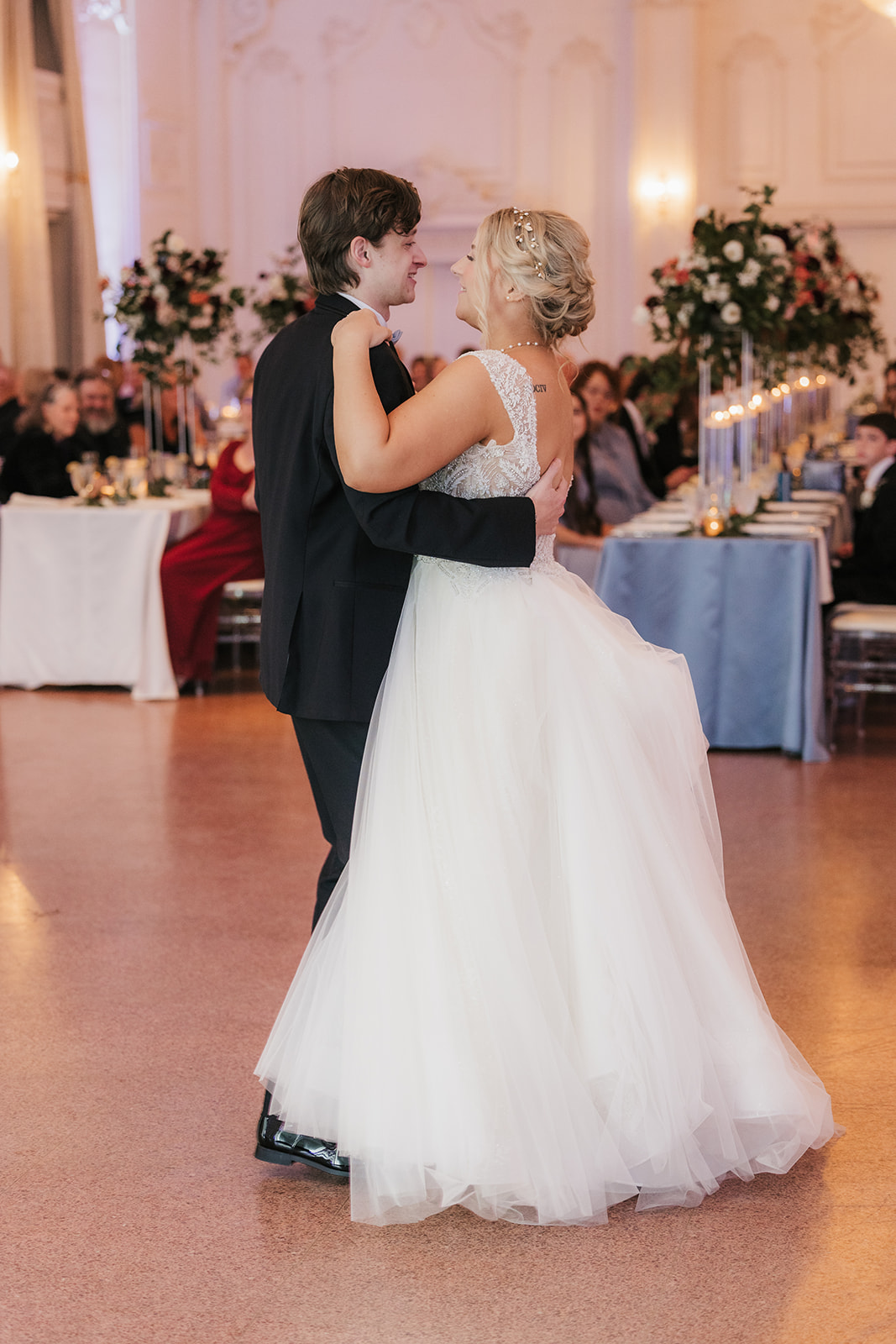 A couple dances together in a formal setting, surrounded by seated guests and floral arrangements.