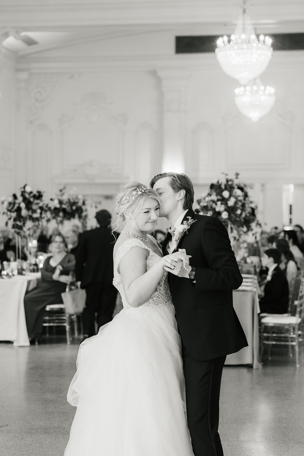 A couple dances together in a formal setting, surrounded by seated guests and floral arrangements.