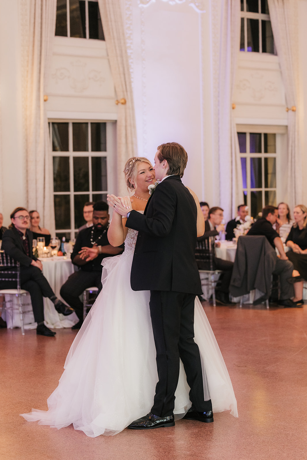 A couple dances together in a formal setting, surrounded by seated guests and floral arrangements.