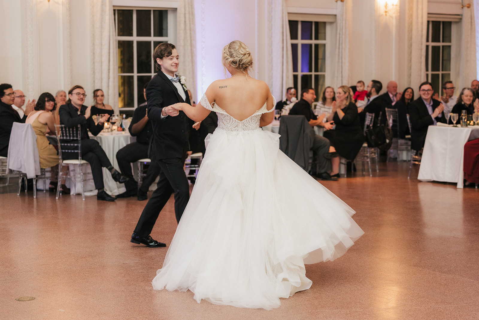 A couple dances together in a formal setting, surrounded by seated guests and floral arrangements.