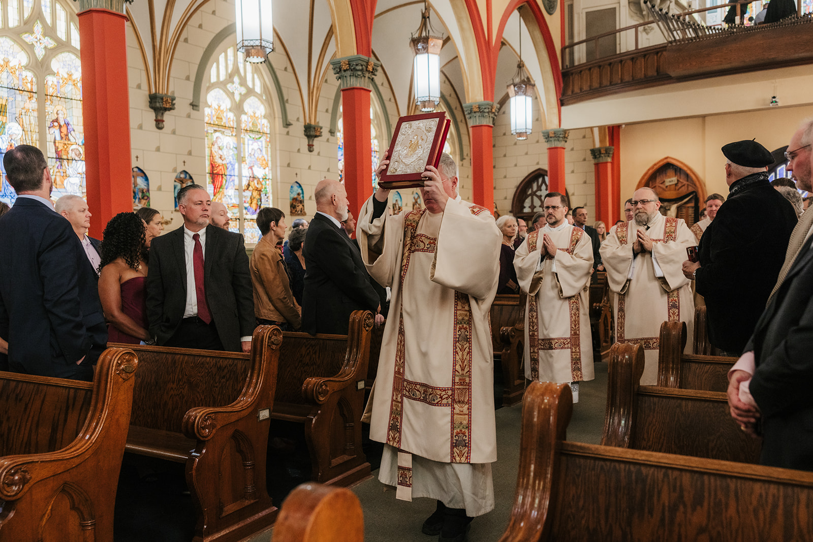 A religious procession in a church, with a leader holding an ornate book, followed by clergy members. Parishioners are seated in wooden pews. Wedding ceremony at the Holy family cathedral in Tulsa