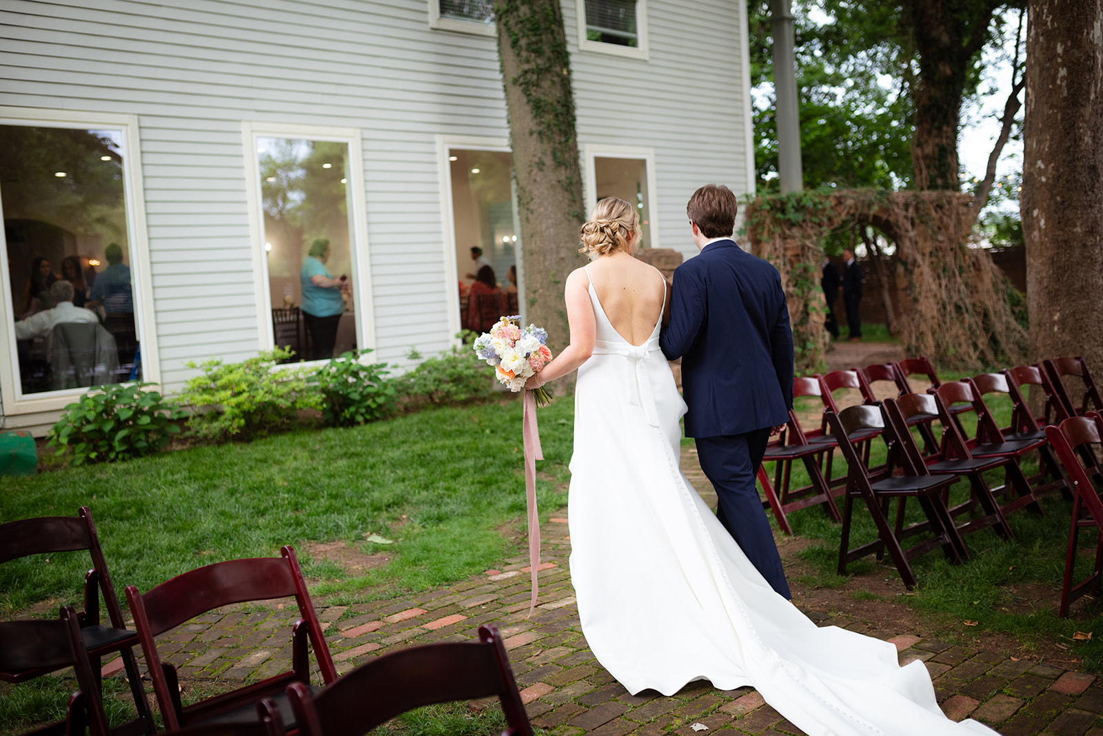 A bride and groom walk down an outdoor aisle lined with empty chairs. The bride wears a white dress with a long train, and the groom is in a dark suit. Trees and a white building are in the background.