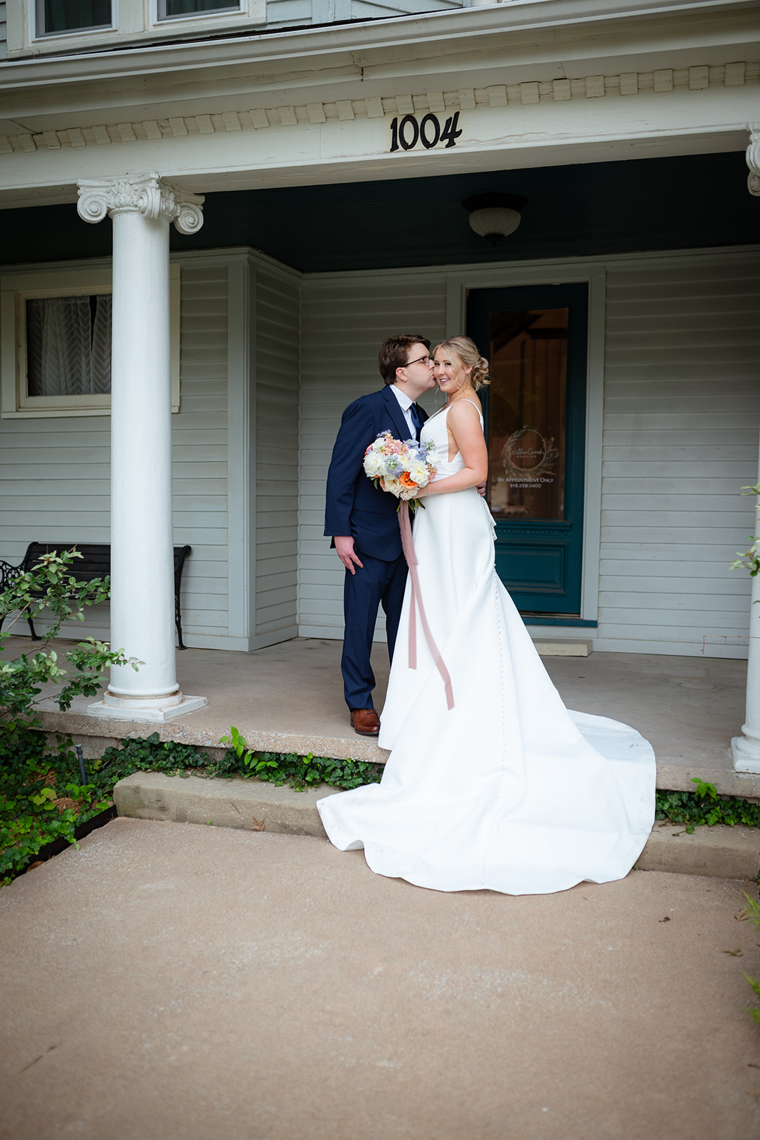 A bride and groom stand together in front of a large, two-story house with columns. The bride holds a bouquet.