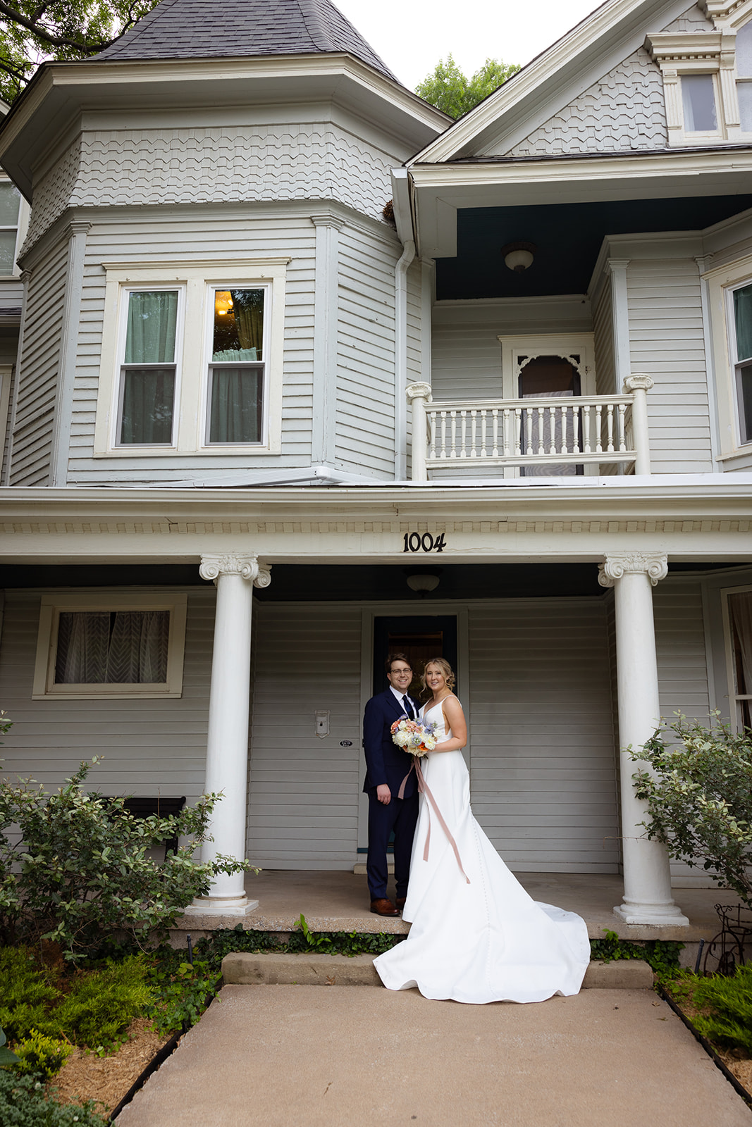 A bride and groom stand together in front of a large, two-story house with columns. The bride holds a bouquet.