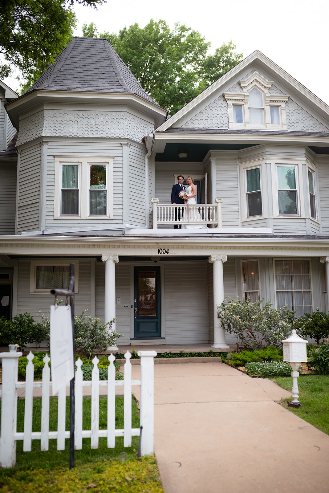 A bride and groom stand together in front of a large, two-story house with columns. The bride holds a bouquet.