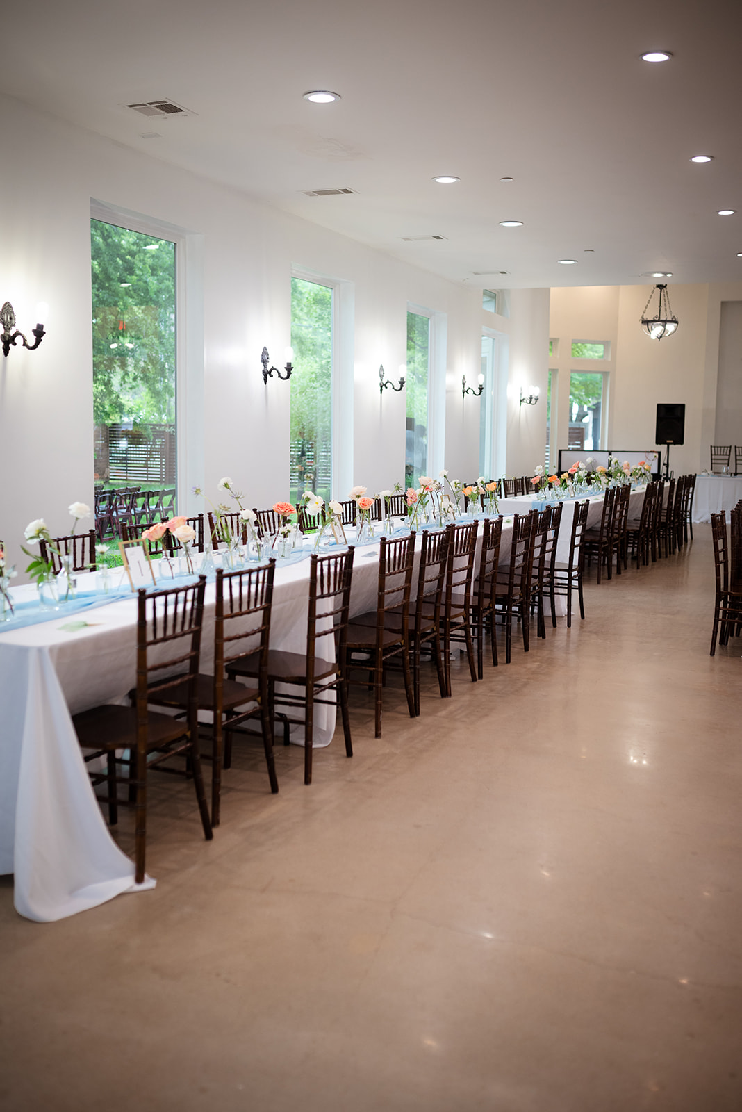 A long, elegant dining table set for a formal event, with white tablecloths, chairs, and floral centerpieces in a brightly lit room with large windows.