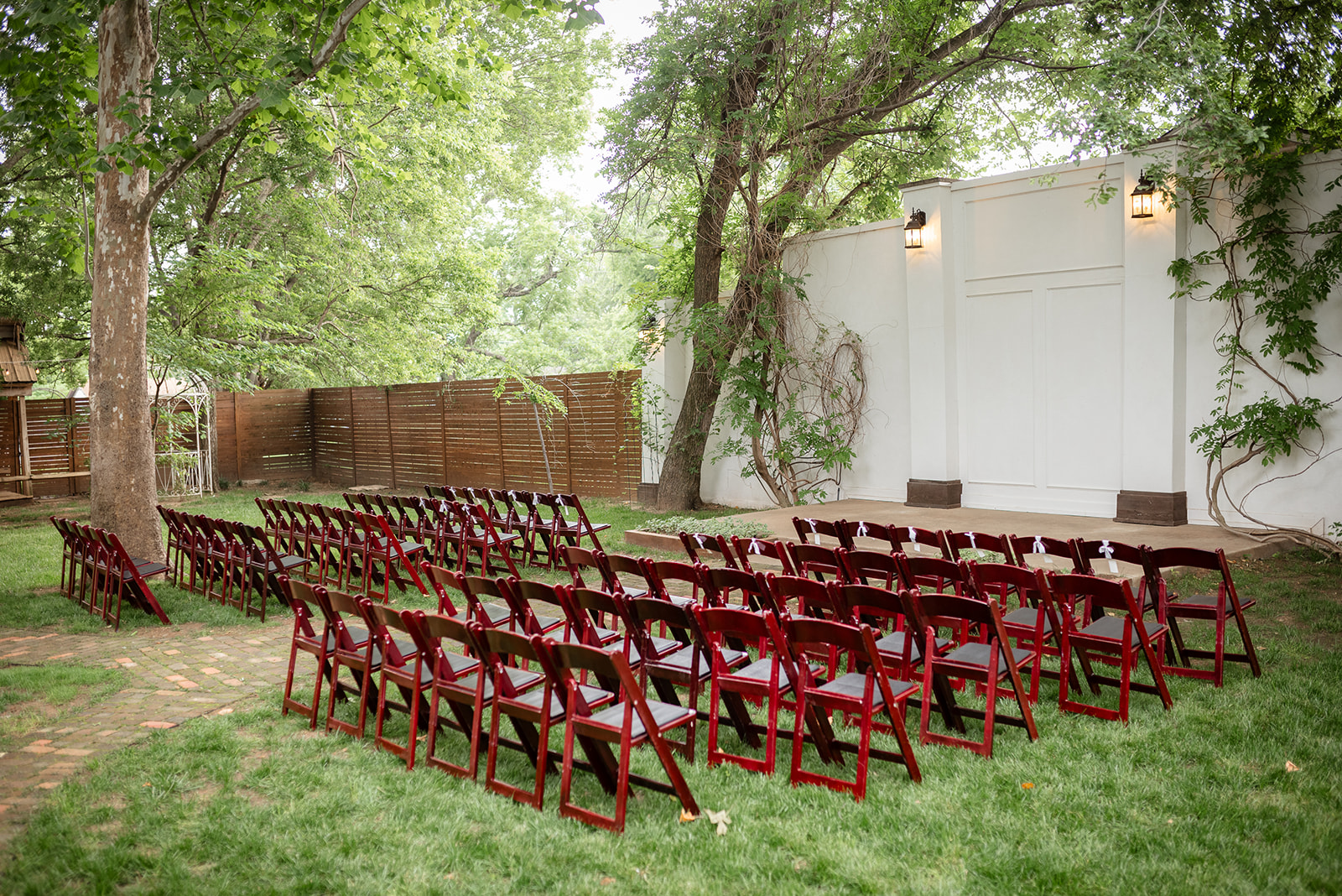 Outdoor wedding setup with rows of wooden chairs facing a small stage surrounded by trees and greenery.