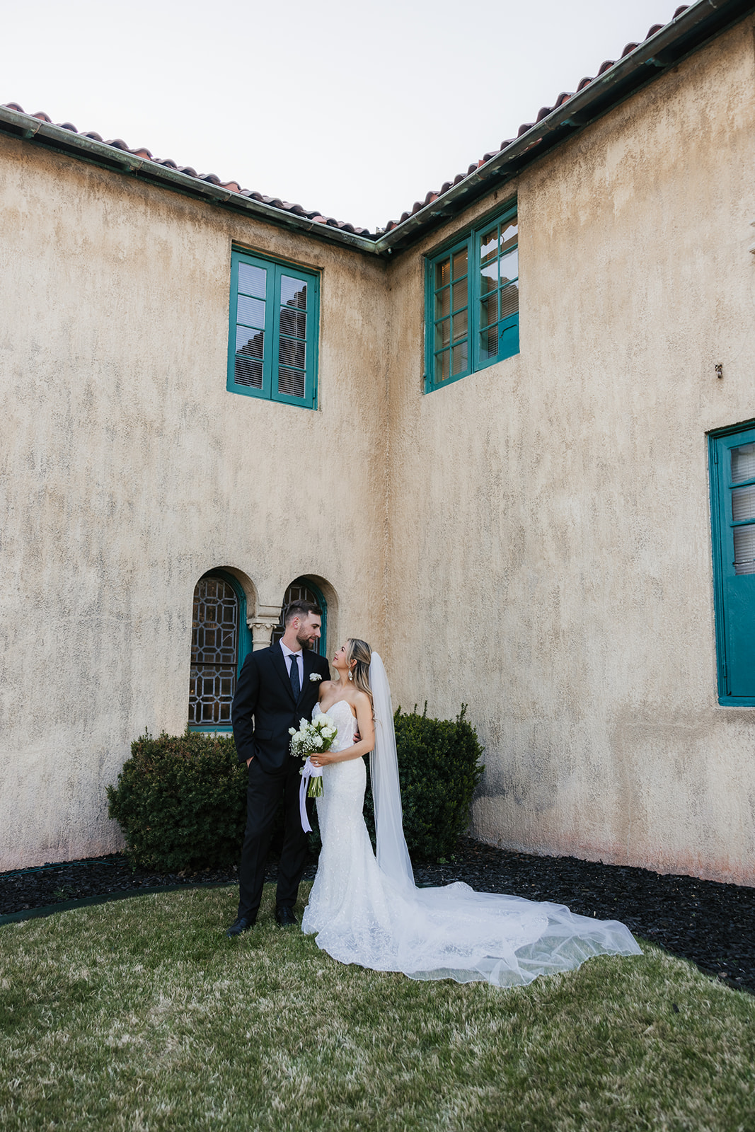 Bride and groom standing on steps of a historic building with blue doors and windows, flanked by tall potted plants. A modern cylindrical structure is visible in the background | Top 6 Historic Mansion Wedding Venues in Tulsa
