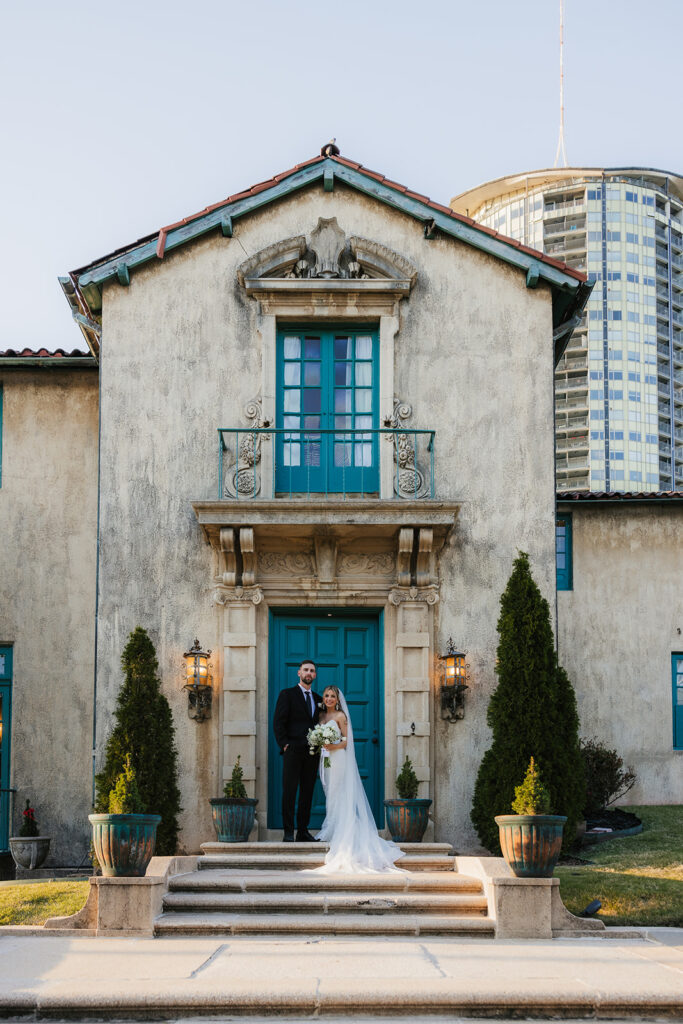 Bride and groom standing on steps of a historic building with blue doors and windows, flanked by tall potted plants. A modern cylindrical structure is visible in the background | Top 6 Historic Mansion Wedding Venues in Tulsa